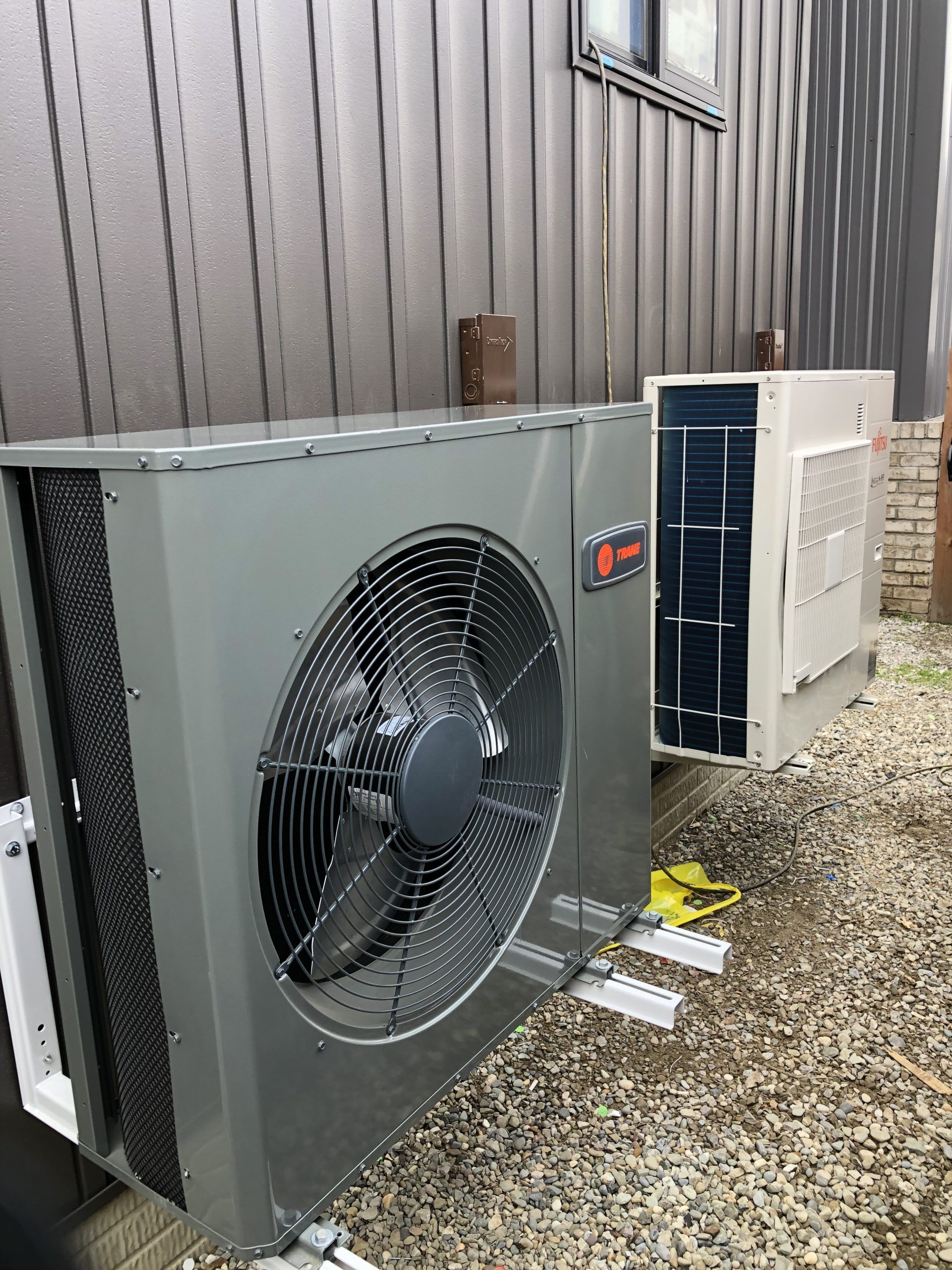Two gray and white HVAC units mounted on a building's exterior, near gravel.