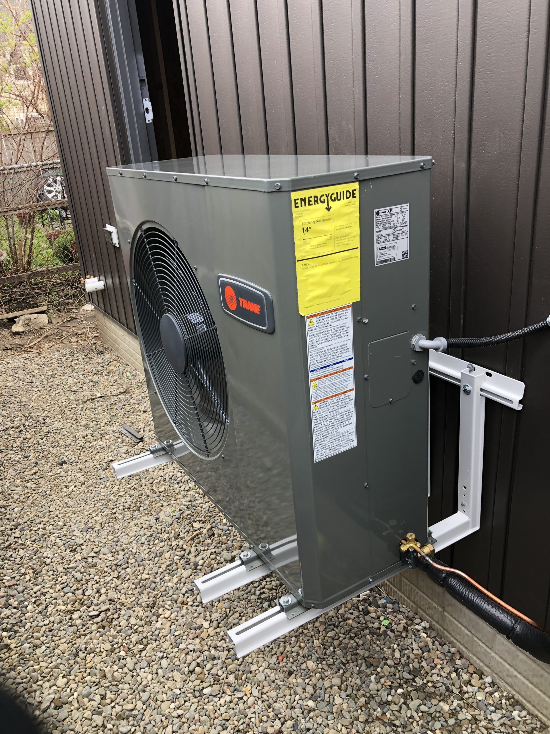 Gray Trane heat pump unit mounted on a brown wall with aluminum brackets, outdoors.