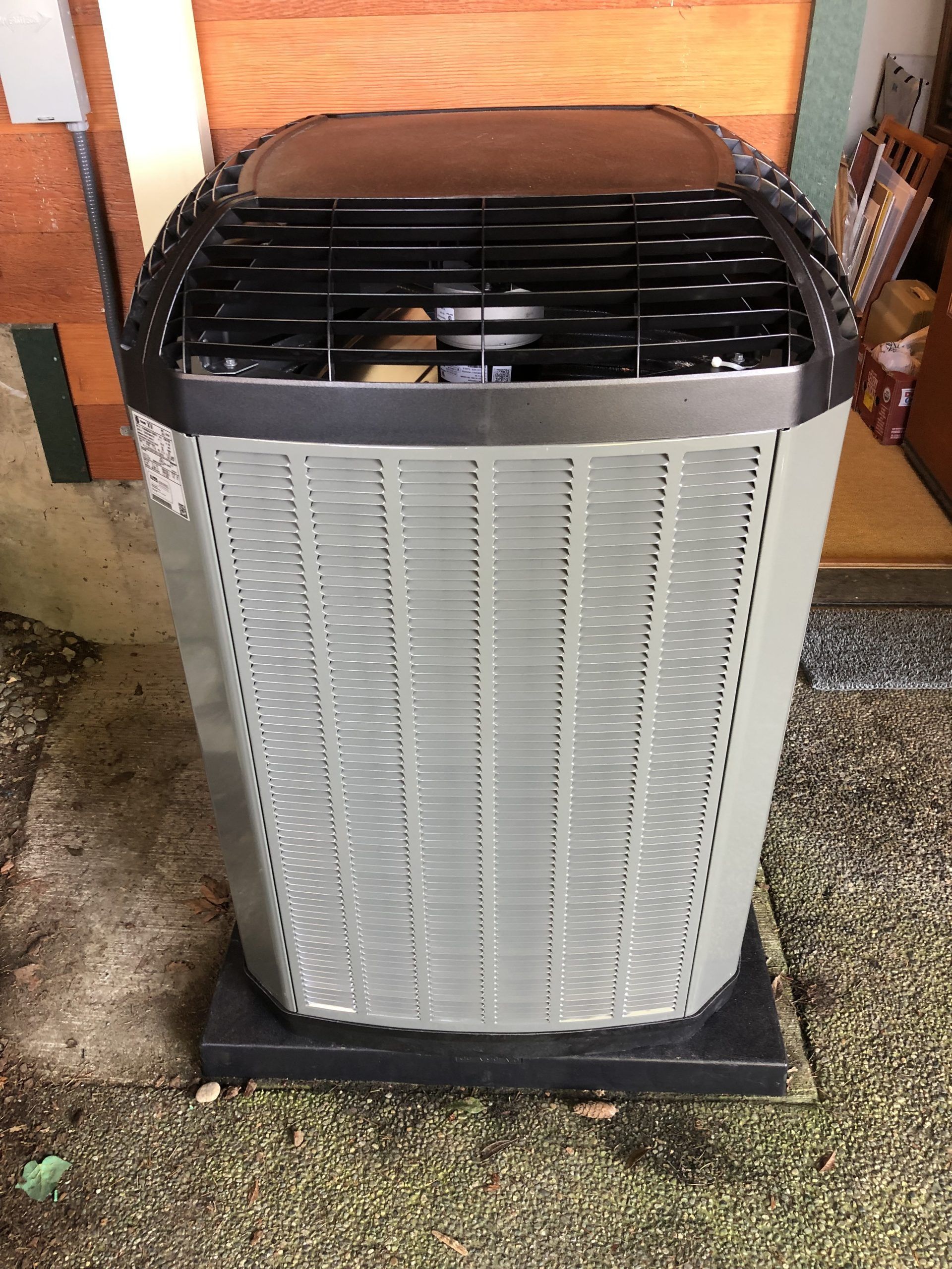 Gray and black air conditioner unit on a black base, set against a wooden wall and gravel.