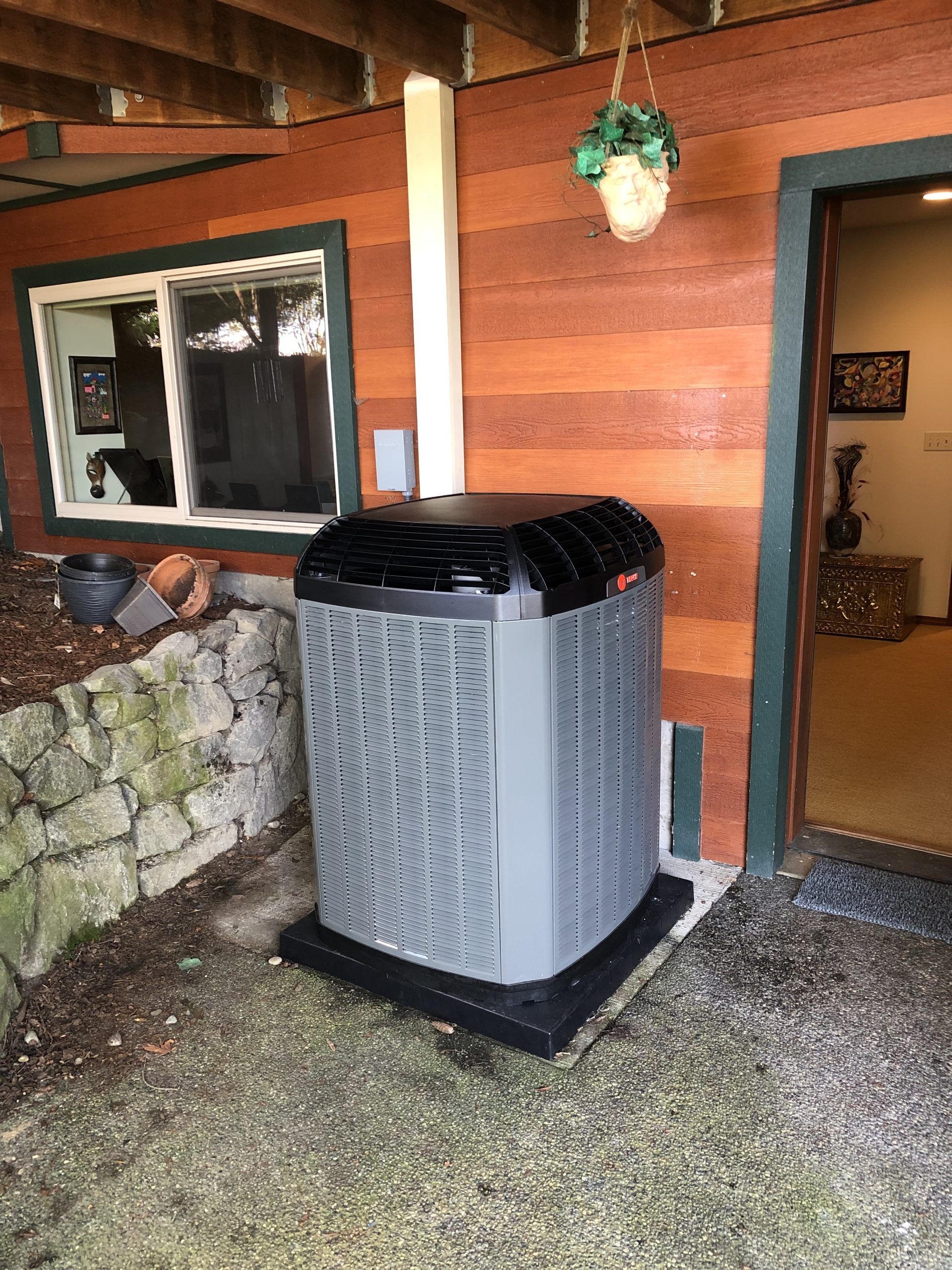 Air conditioner unit outside a building with brown siding and a stone wall.