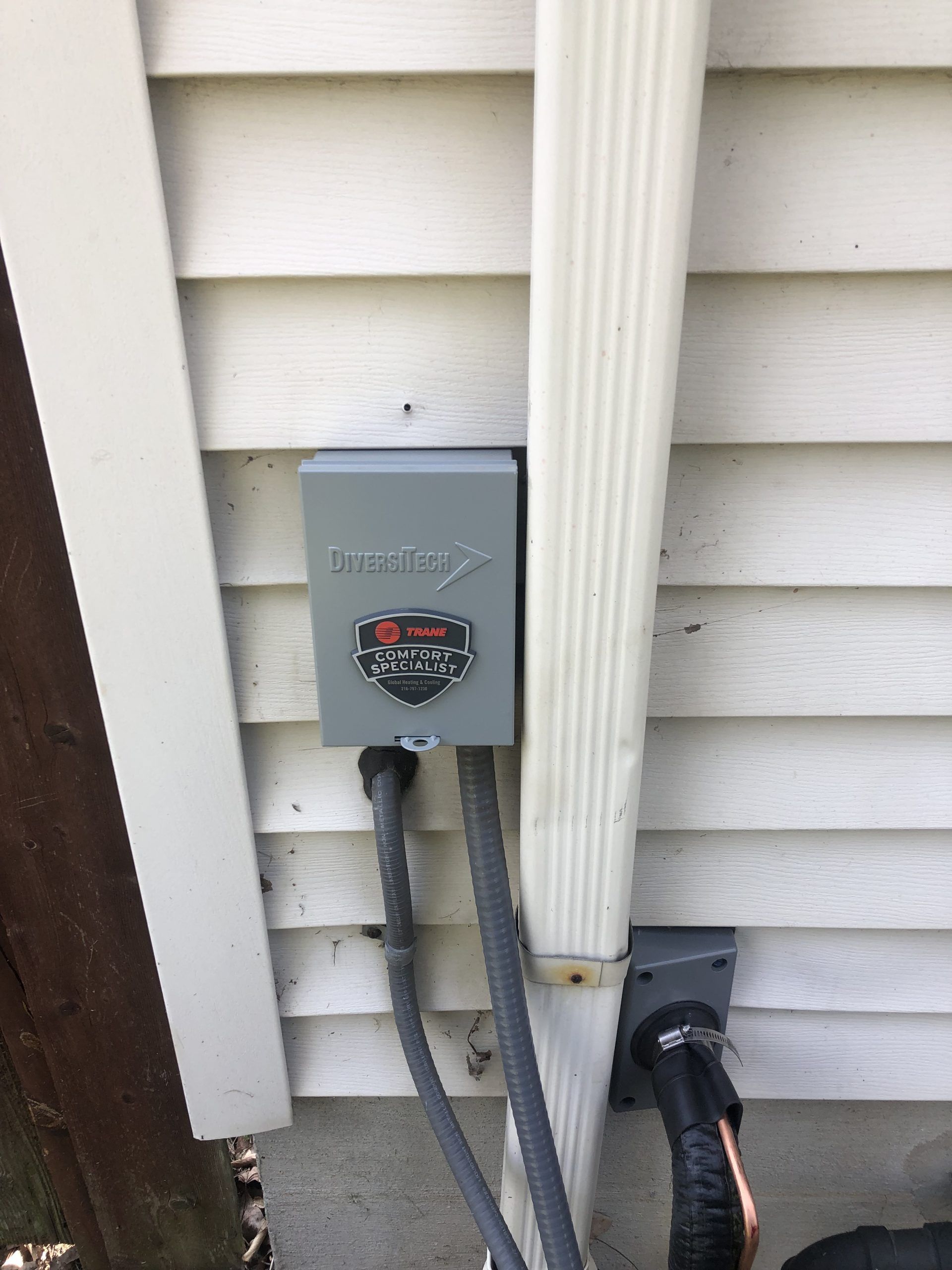 Gray electrical box mounted on white siding next to a downspout and two black wires.