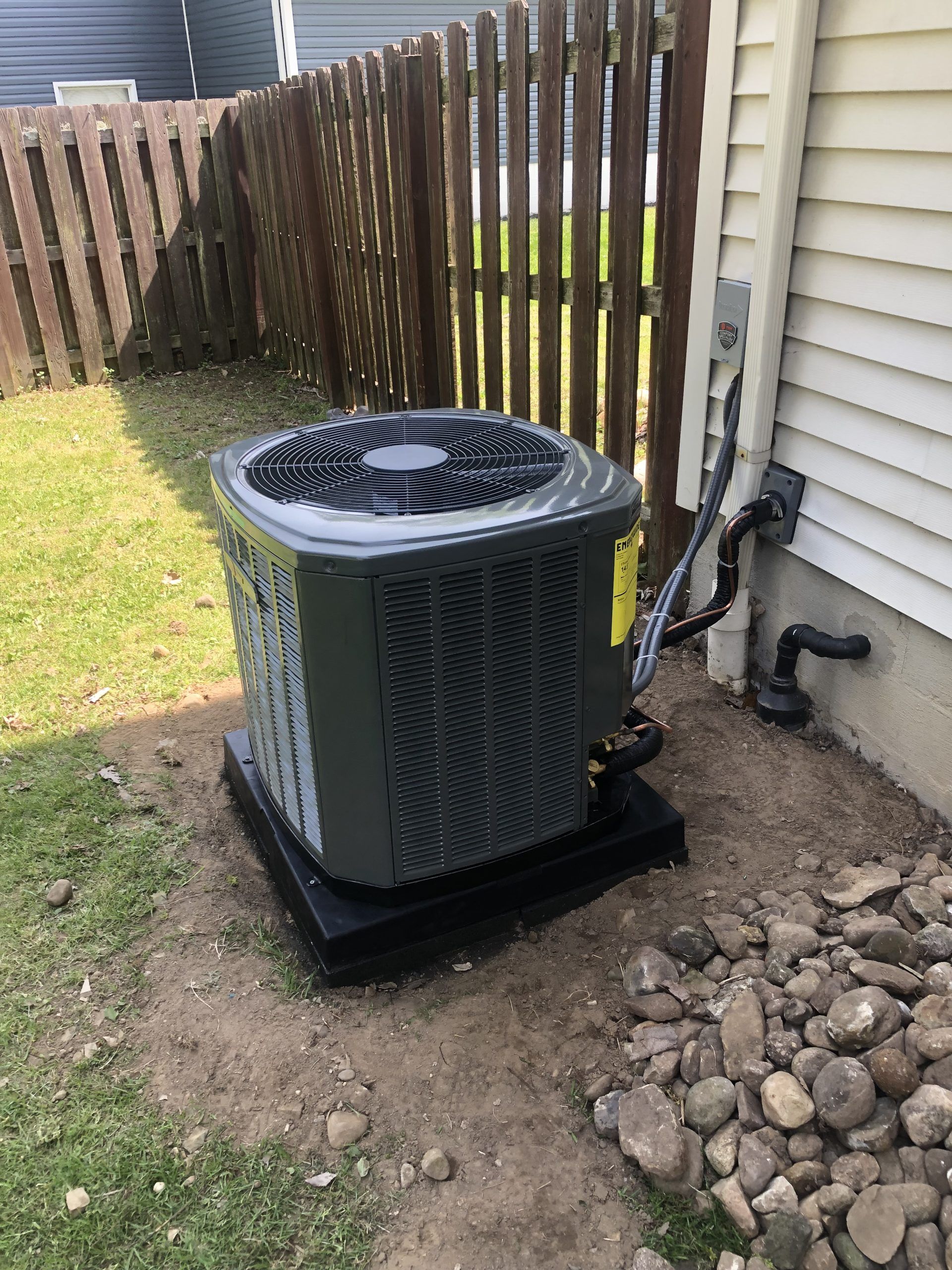 Air conditioning unit outside a house, on a black pad, beside a wooden fence.