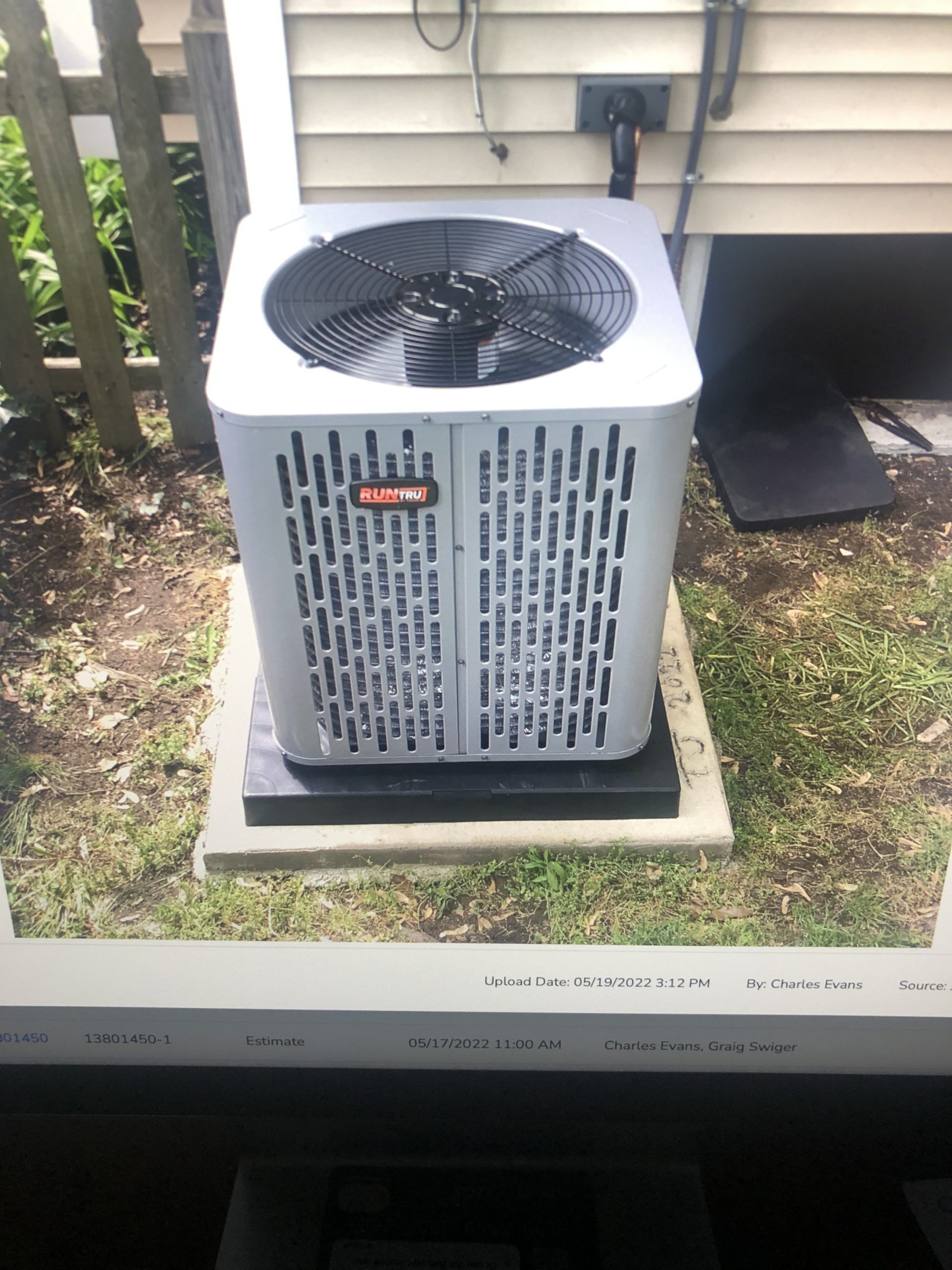 Air conditioner unit, gray, on concrete base, next to a building, outdoors.