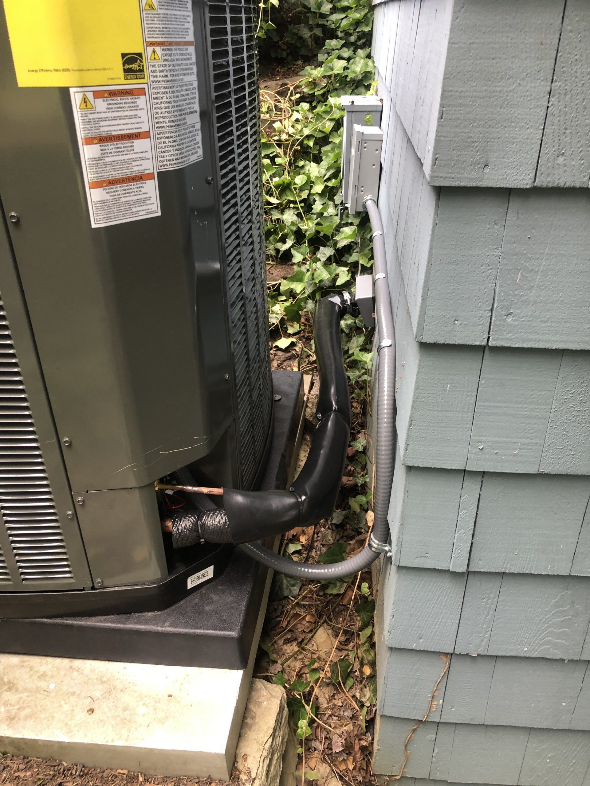 An outdoor air conditioning unit next to a light blue house, with black pipes connecting to it.