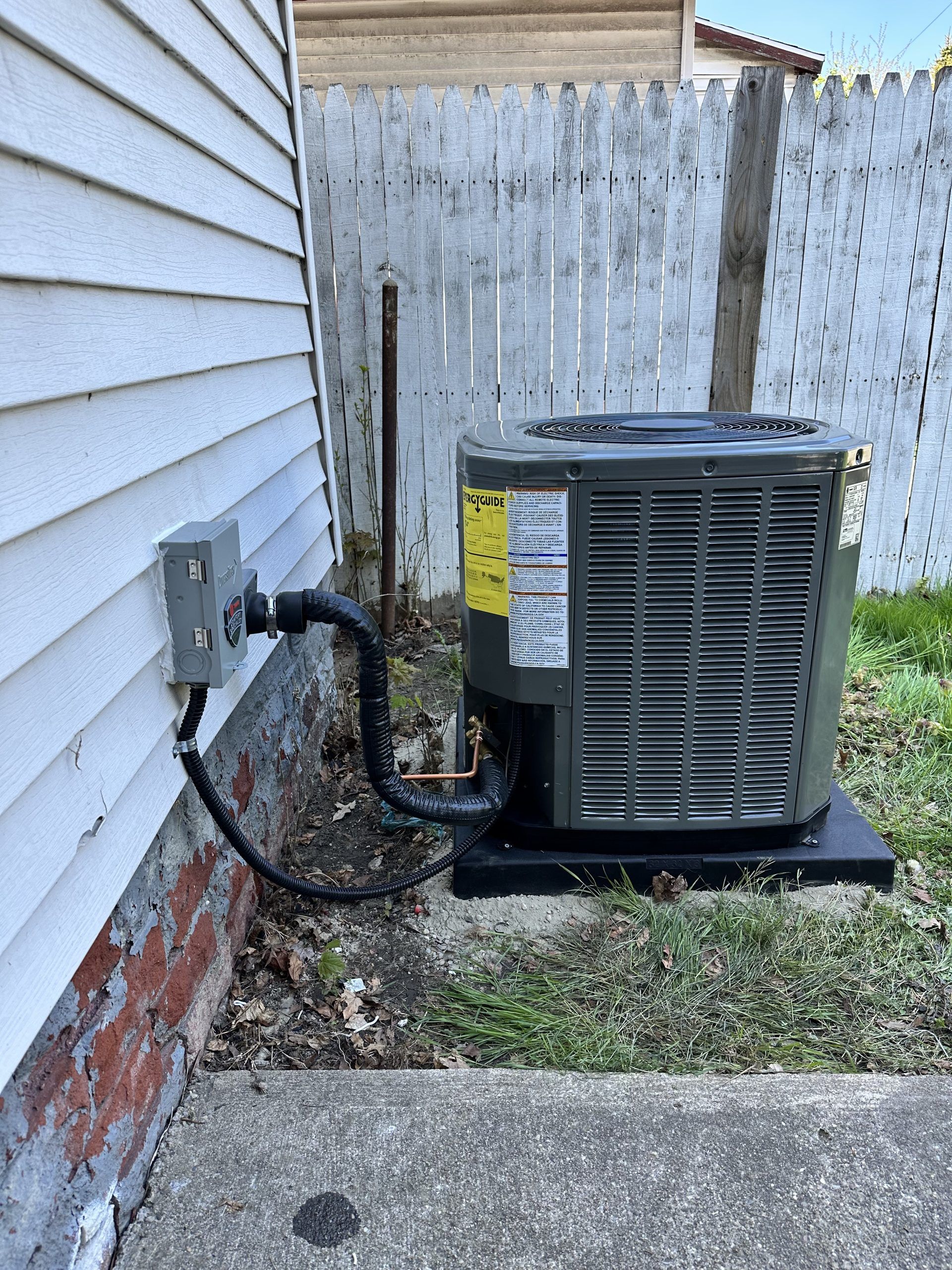 An air conditioning unit next to a house with electrical box and fence.