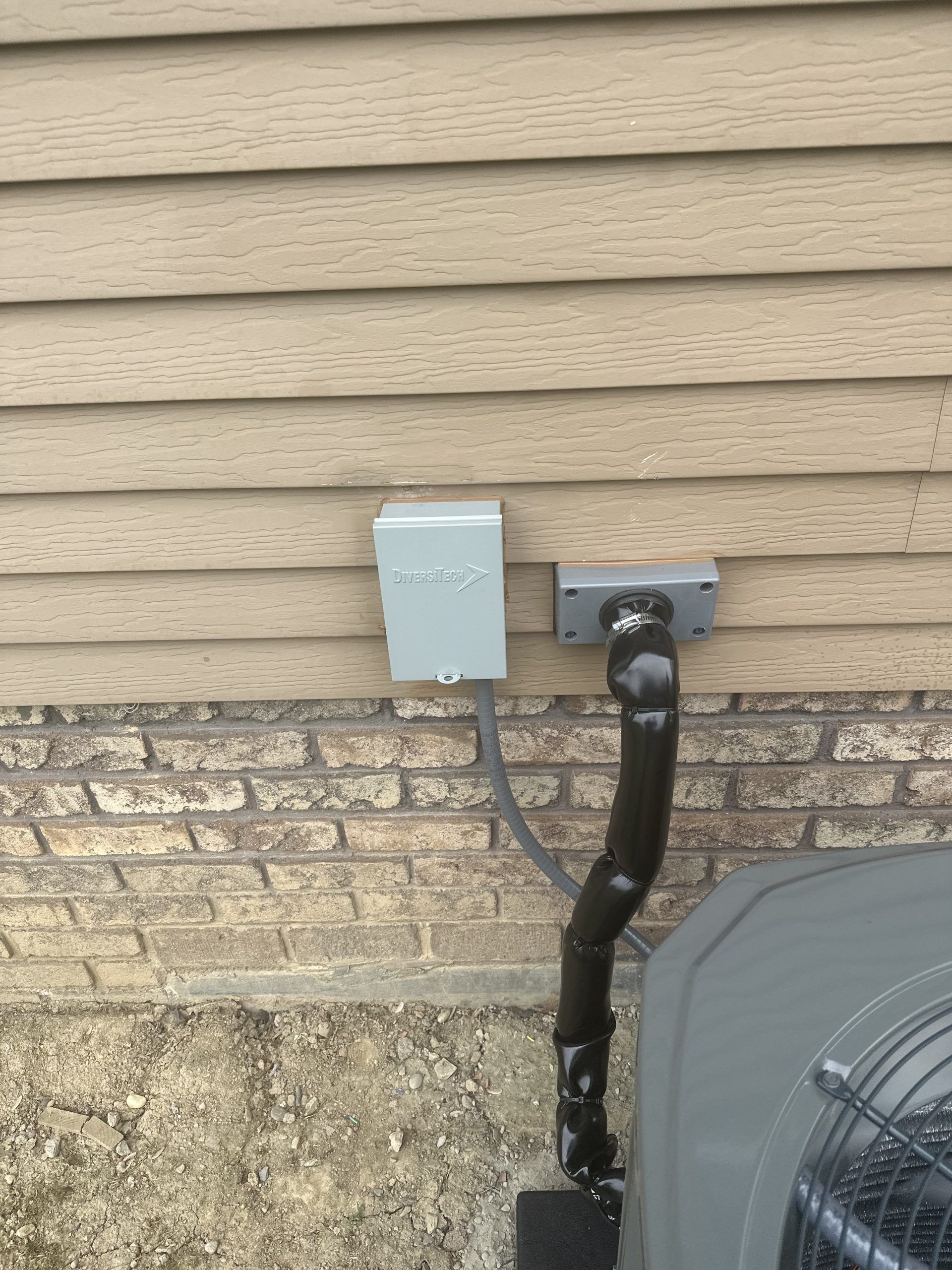 Air conditioner unit with electrical boxes and black pipe on a brick and siding exterior wall.