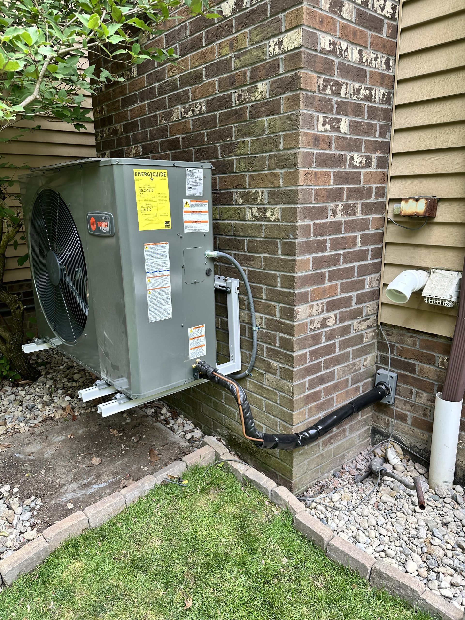 Air conditioning unit mounted on brick, connected to electrical conduit and piping near lawn.