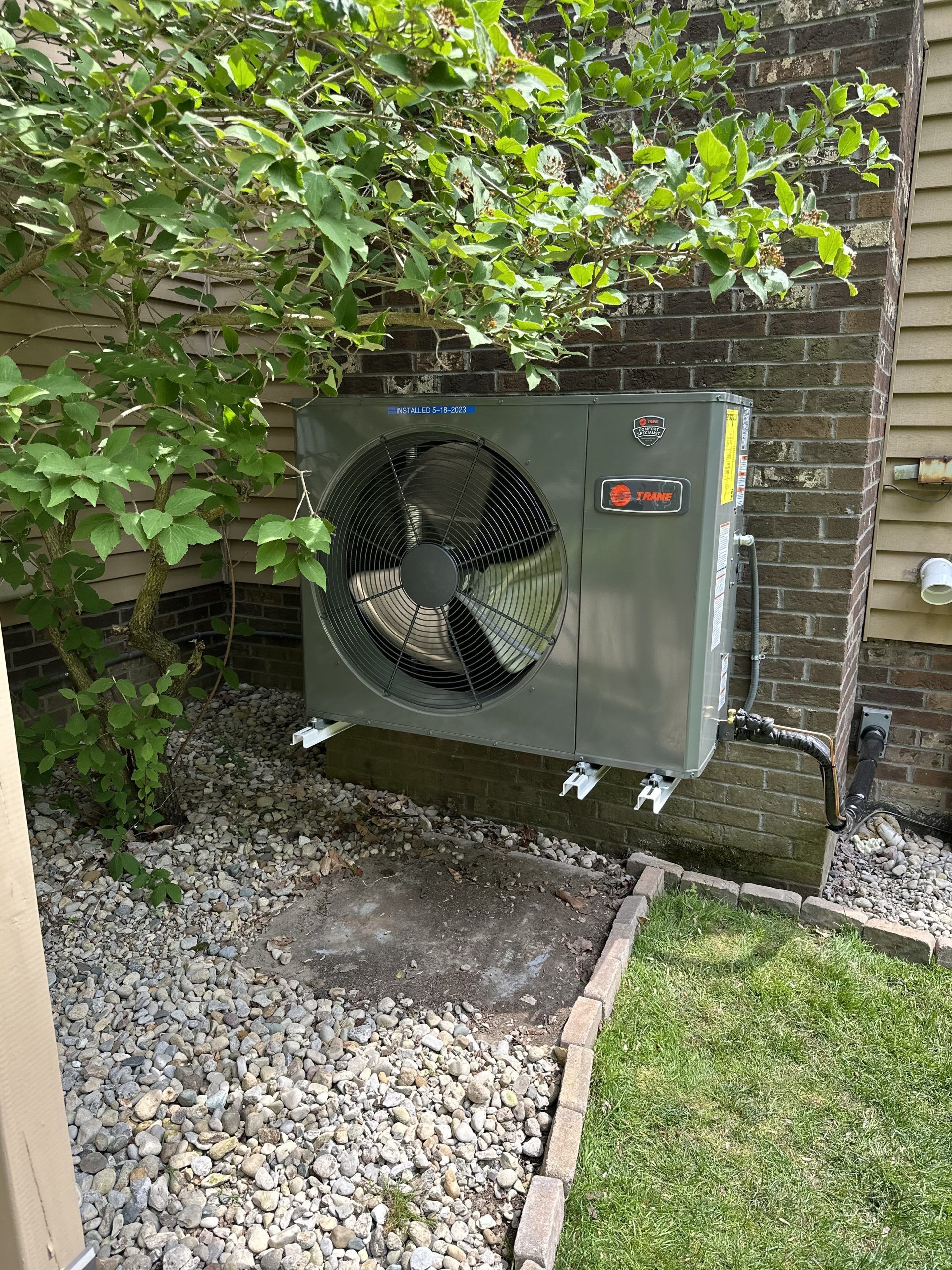 A Trane HVAC unit sits outside against a brick wall, partially shaded by a tree.