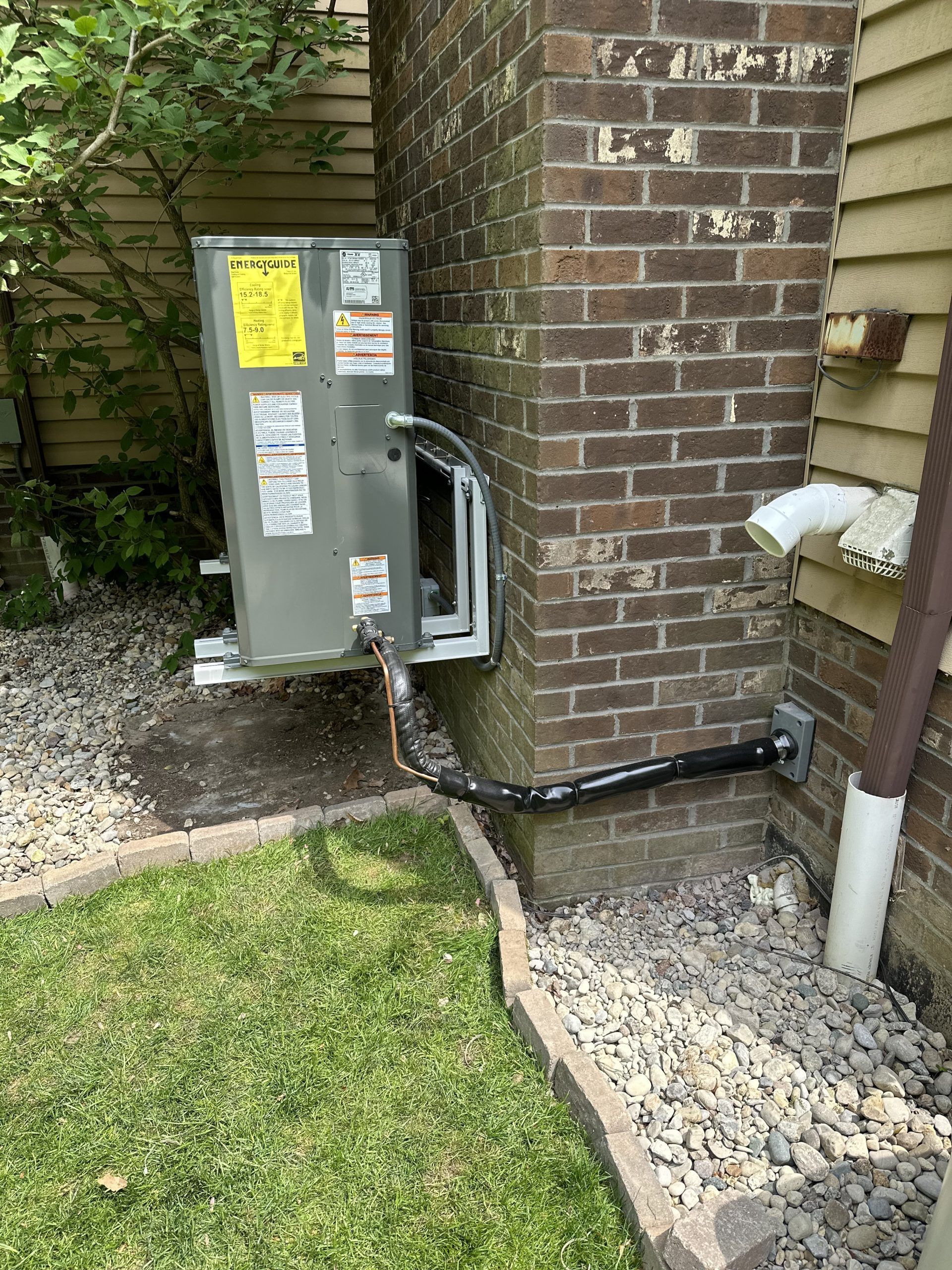 An outdoor HVAC unit next to a brick wall and a house, with black tubing and a white drainpipe.