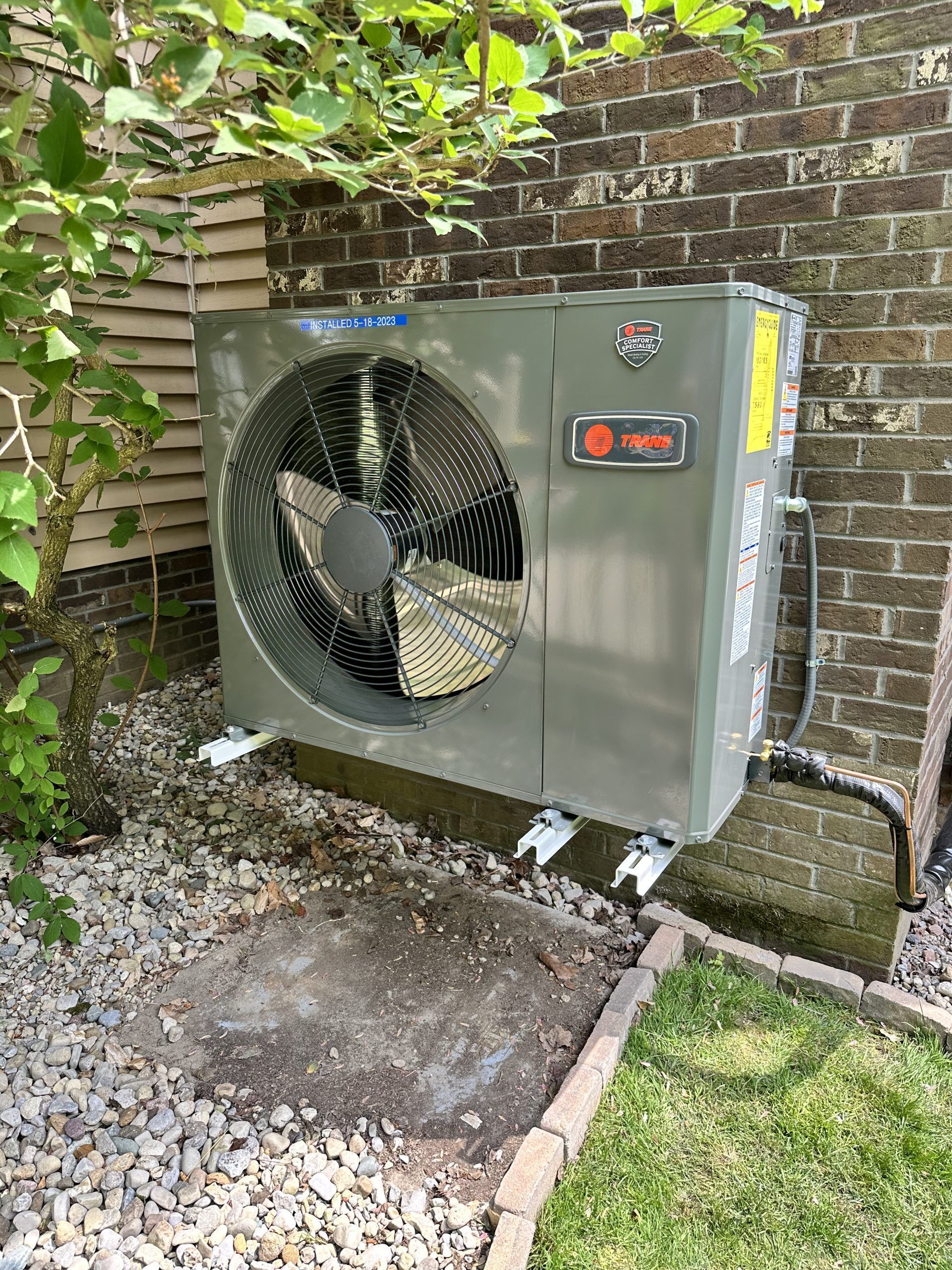 Gray air conditioning unit mounted on a gravel bed near a brick wall and foliage.
