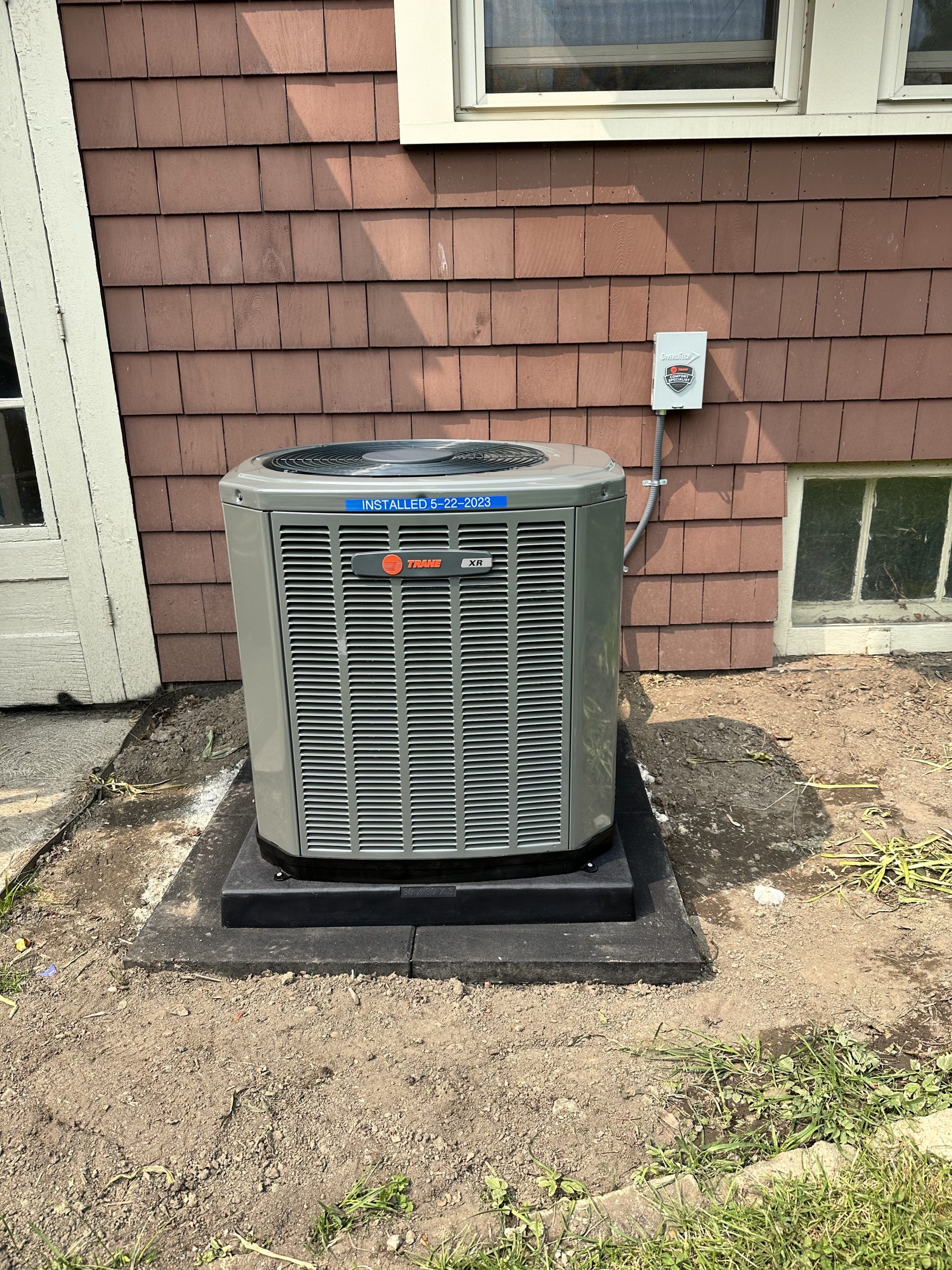 HVAC unit outside a building, set on a dark square base, brown siding, small window, electrical box.