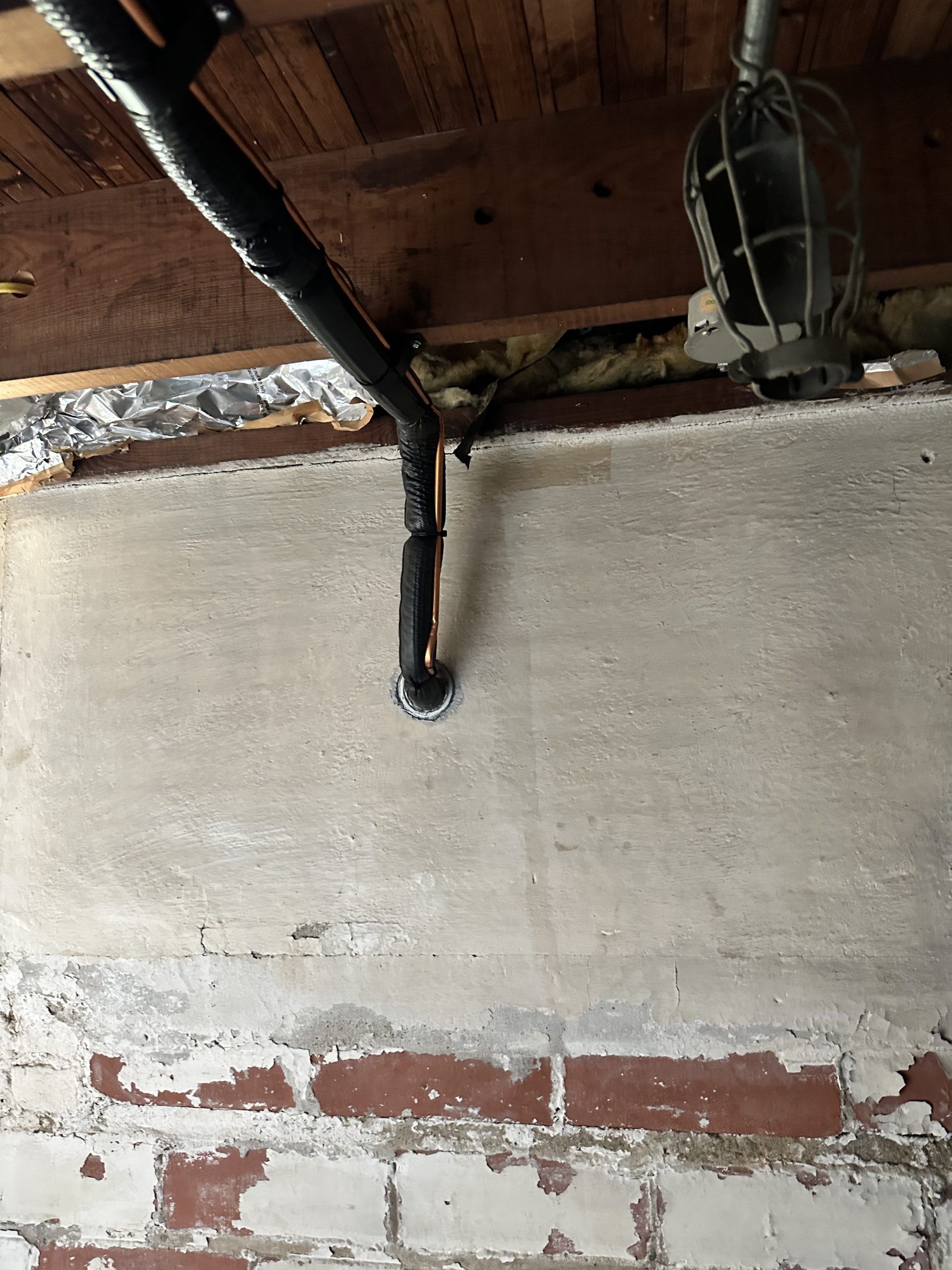 Pipe descending from a wooden ceiling, through a concrete wall, over red bricks.