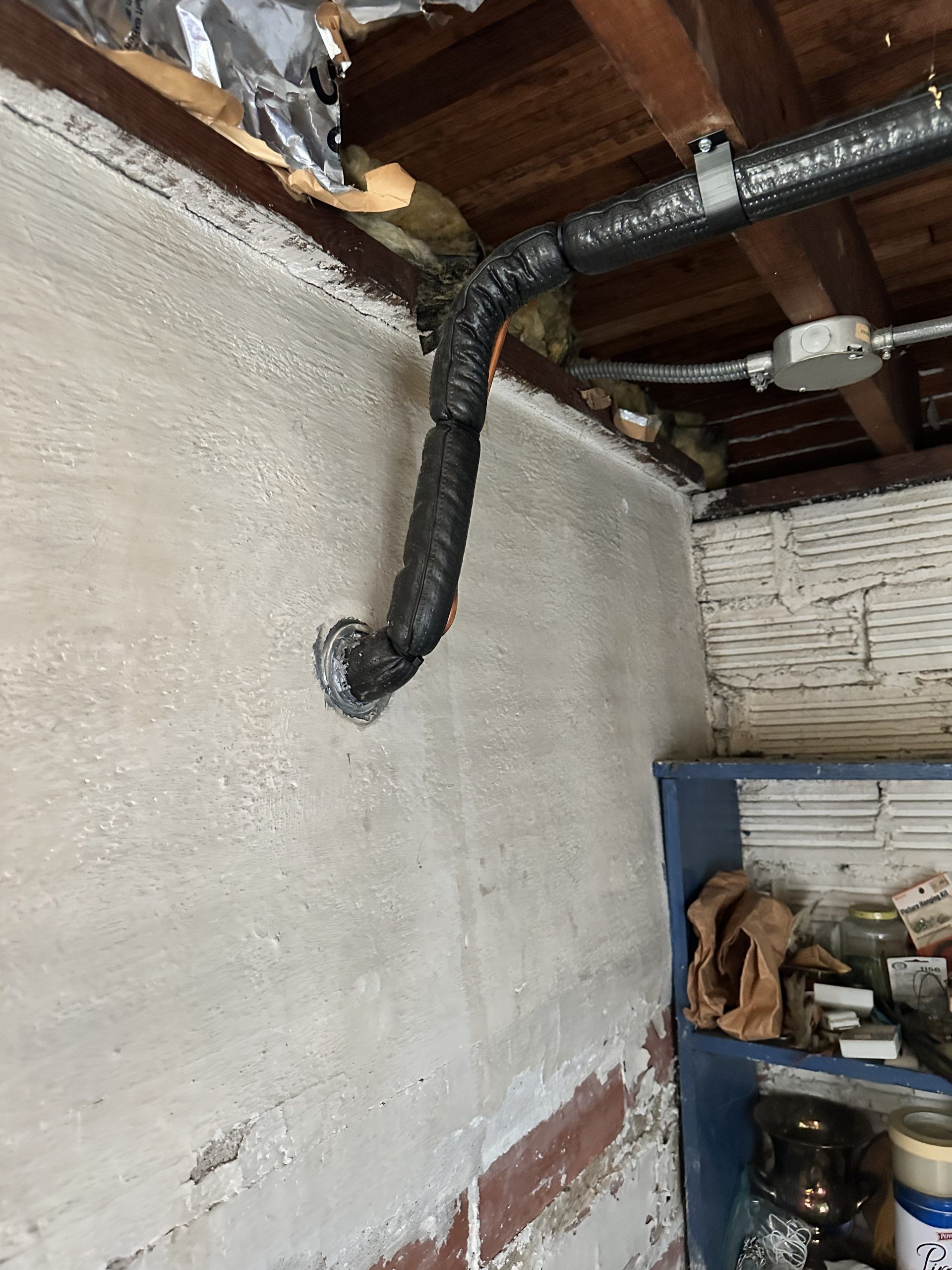 Black vent pipe coming out of a wall with white and gray textured paint. A wooden beam is above.