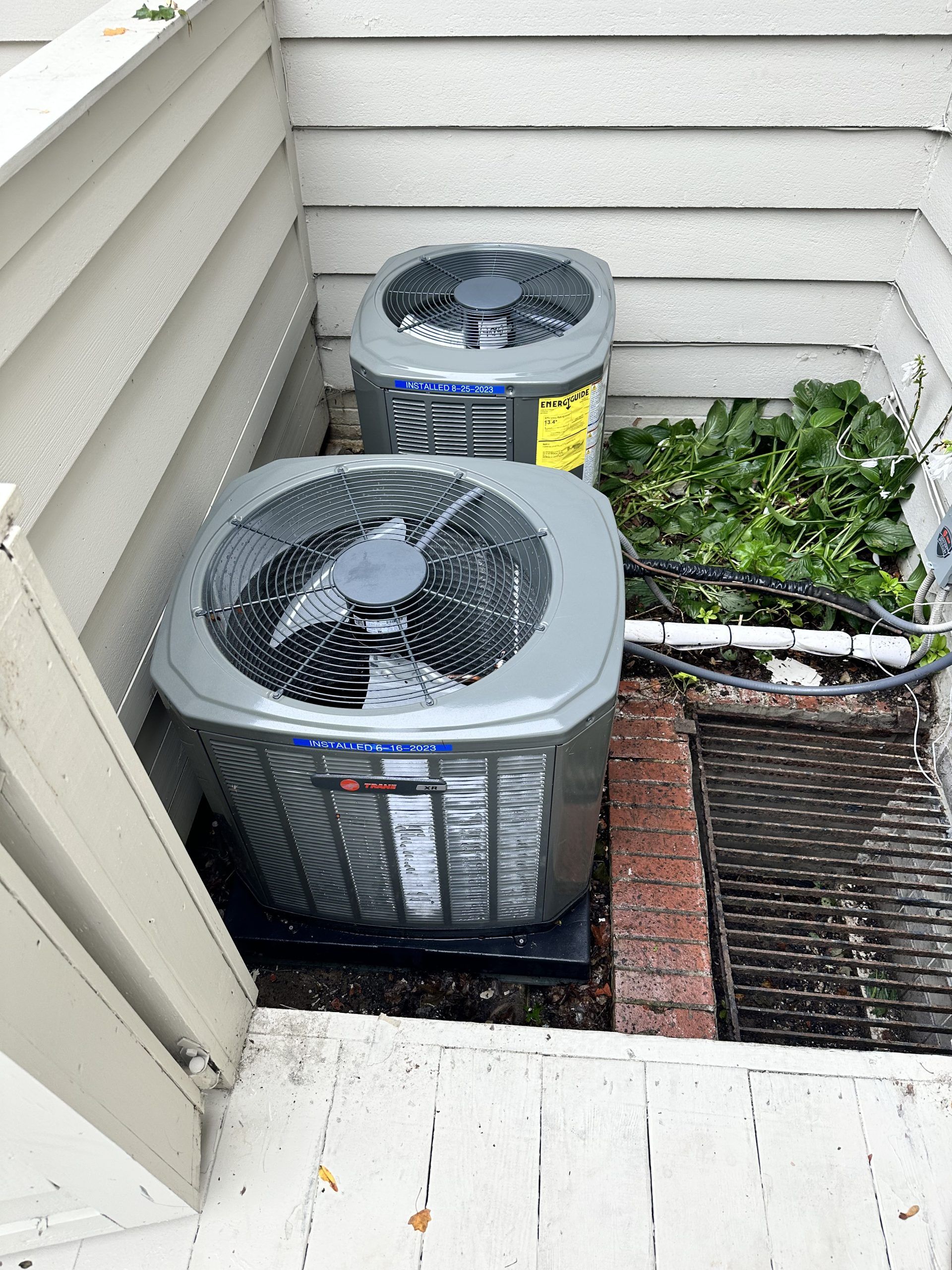 Two outdoor air conditioning units next to greenery in a small, enclosed space. Gray units, white building.