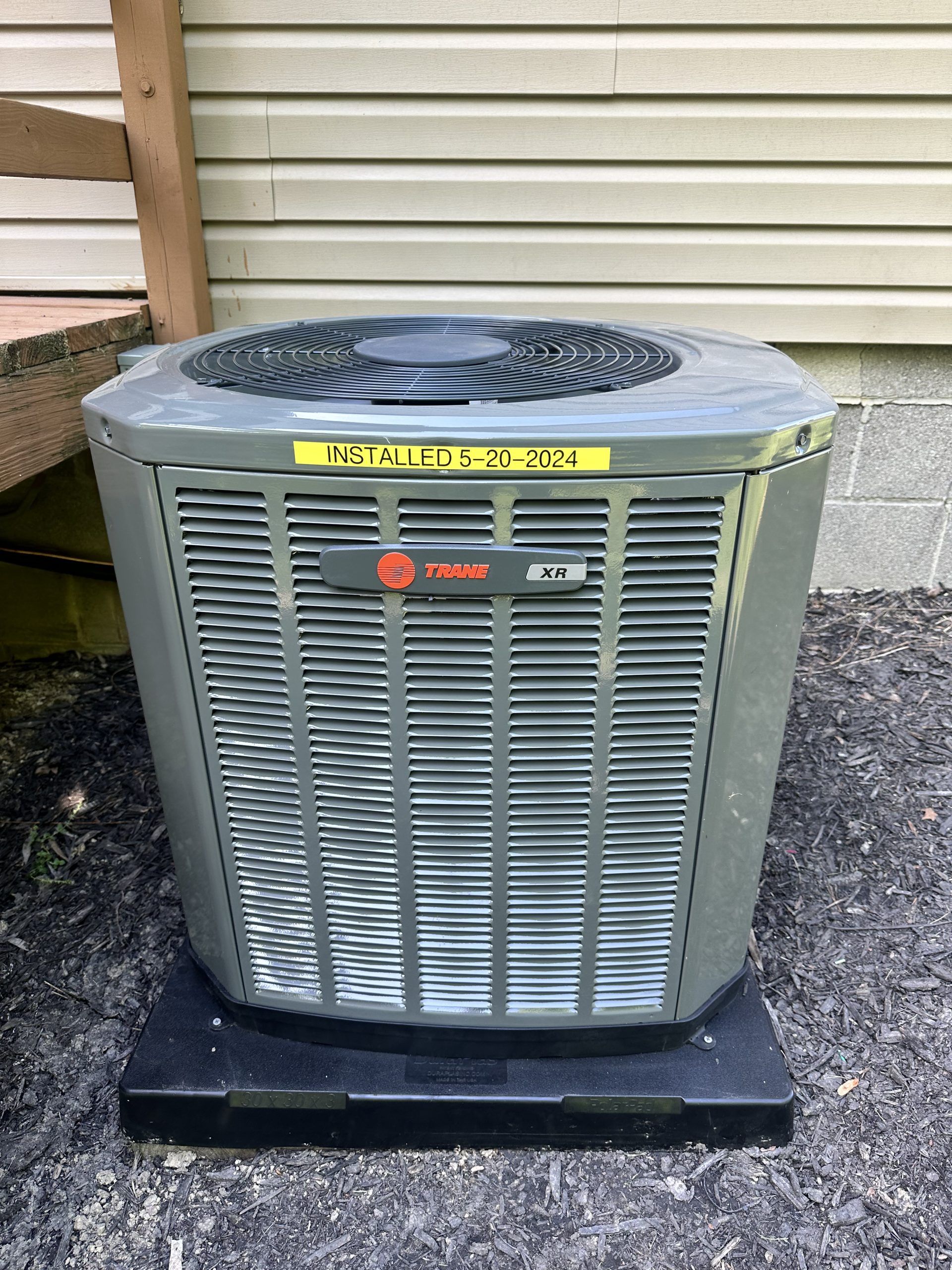 Gray Trane air conditioner unit outdoors, mounted on a black base. Yellow sticker on top.