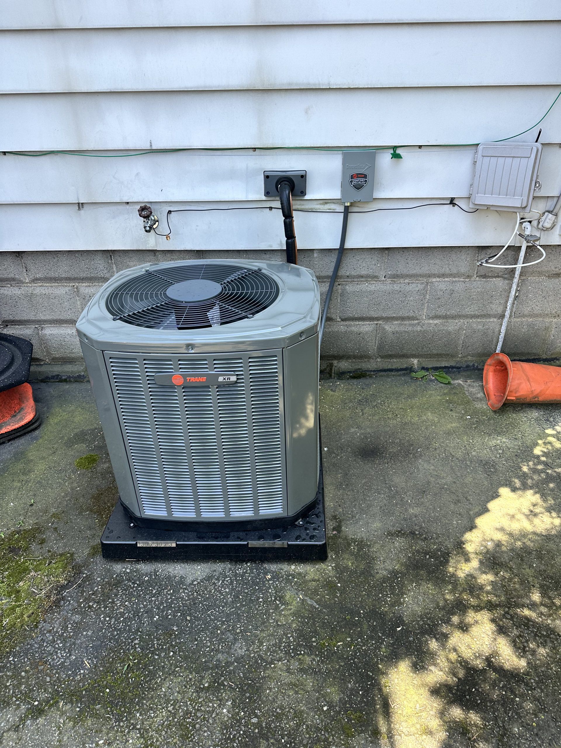 AC unit outdoors, against a white building. Black power cord runs to a nearby electrical box.