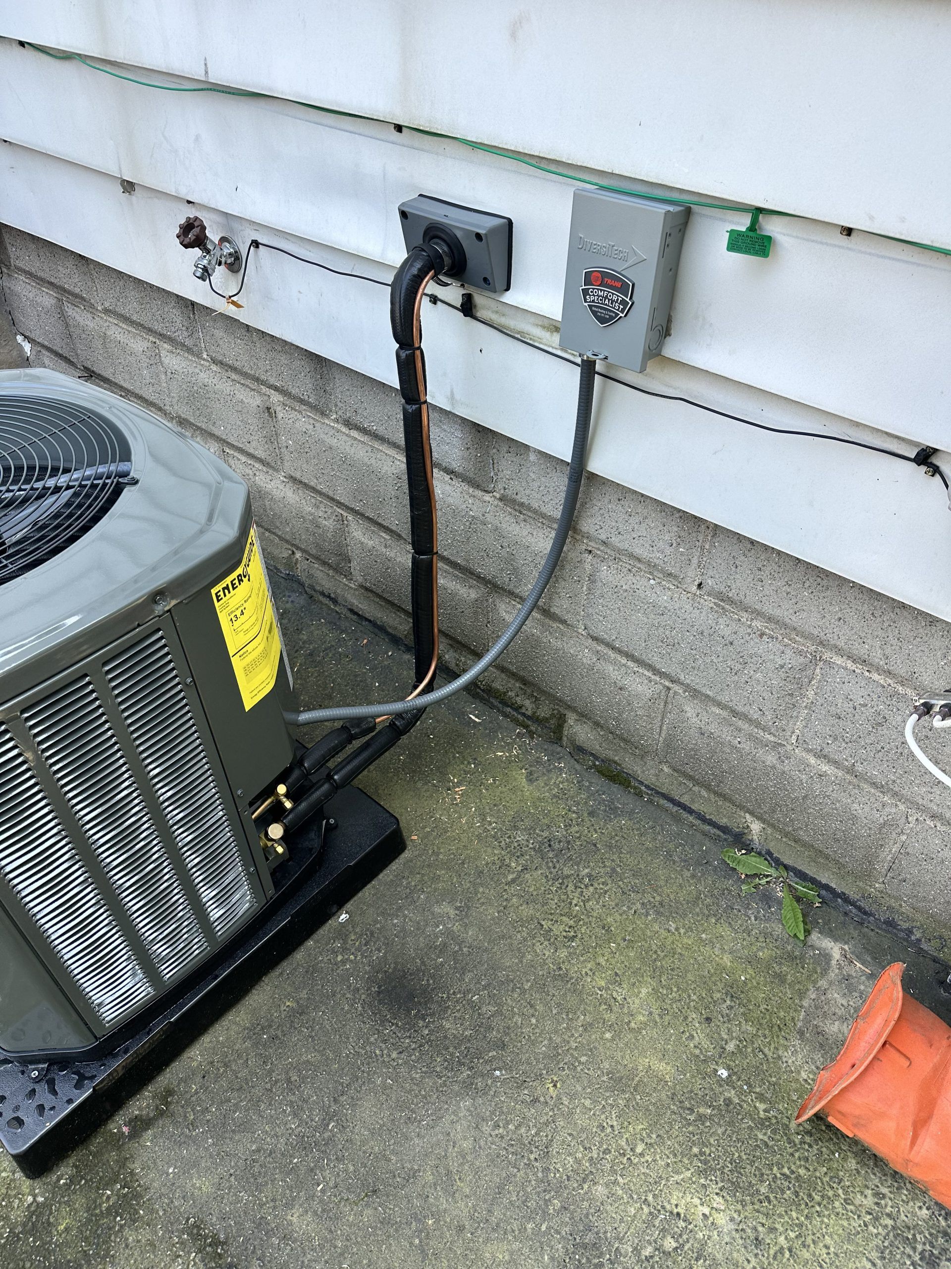 AC unit and electrical box on a building's exterior. Black conduit, gray and white siding, concrete ground.