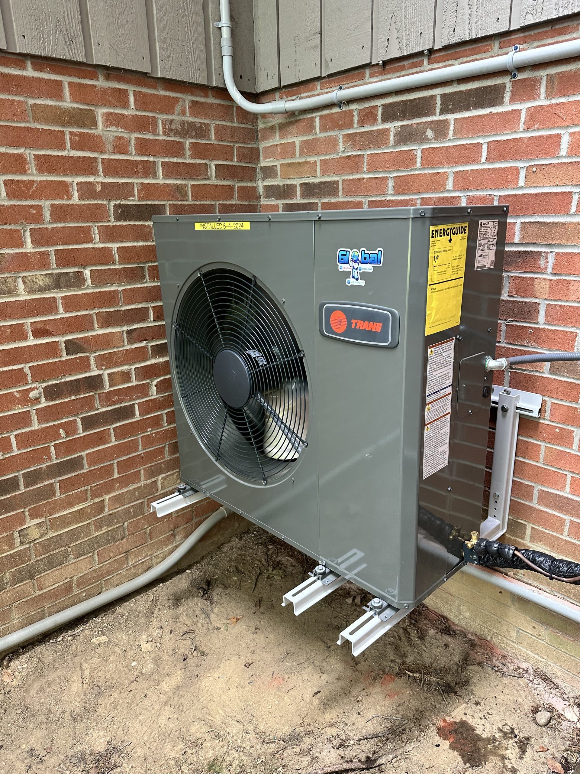 Gray Trane heat pump unit mounted on a brick wall with supporting brackets and piping.