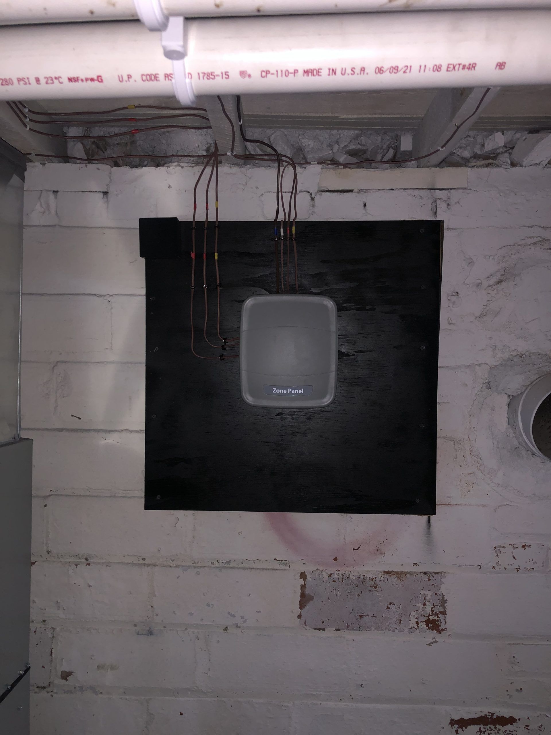A small white device on a black square mounted on a white cinder block wall in a basement, with copper pipes above.