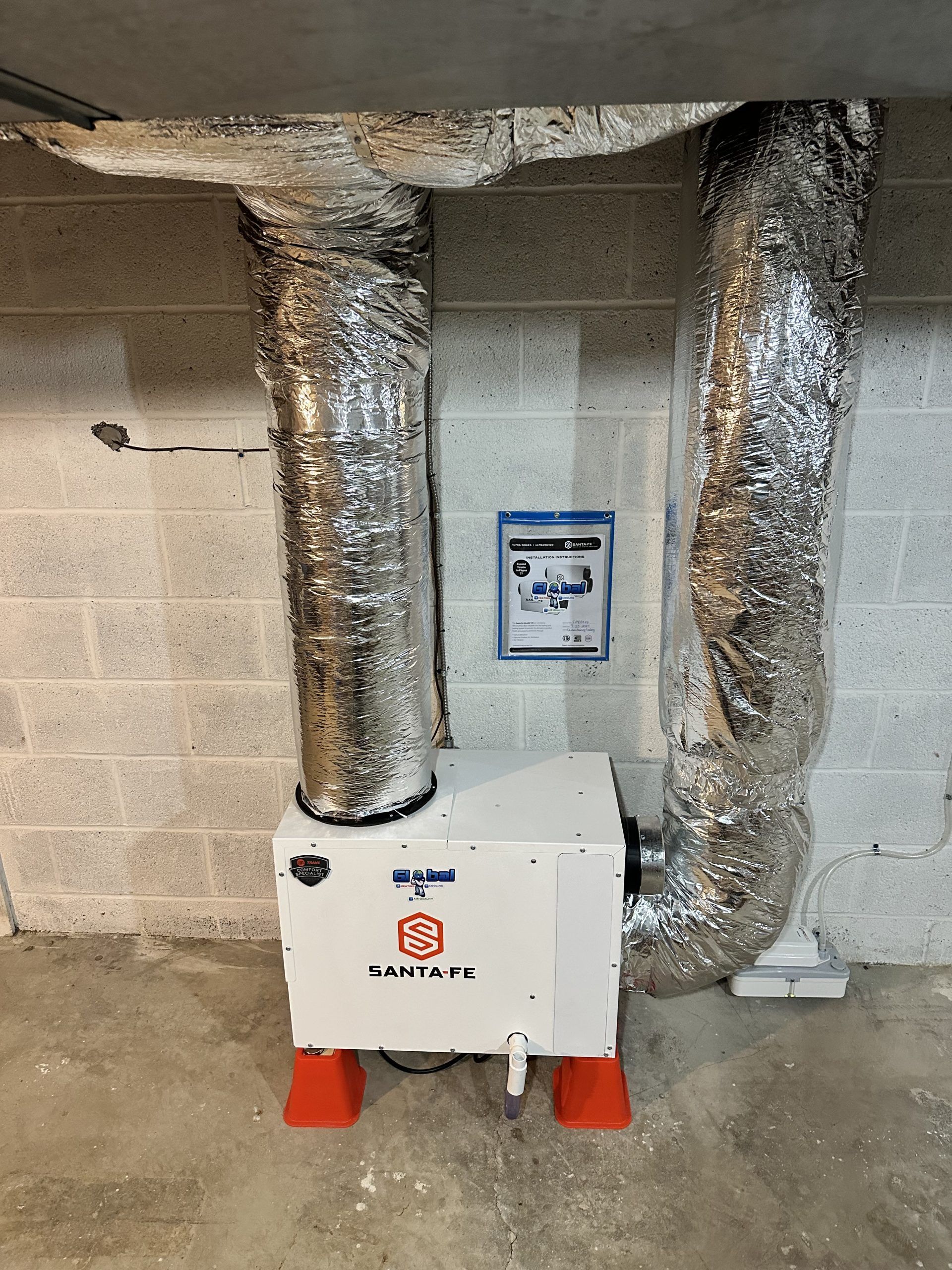White dehumidifier with silver ductwork, placed on orange feet against a concrete block wall.
