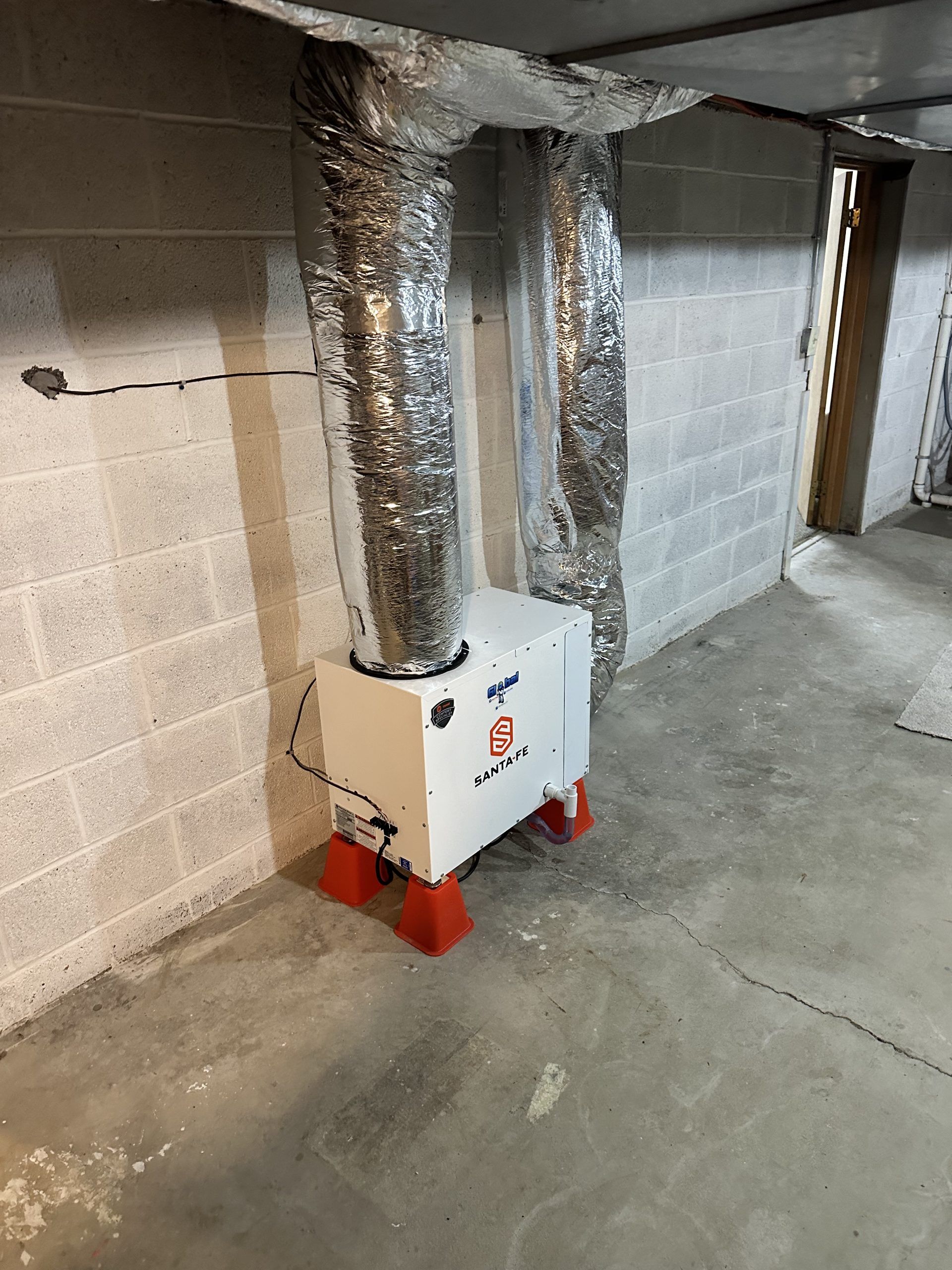 White appliance with red feet, connected to silver ductwork, in a concrete basement.