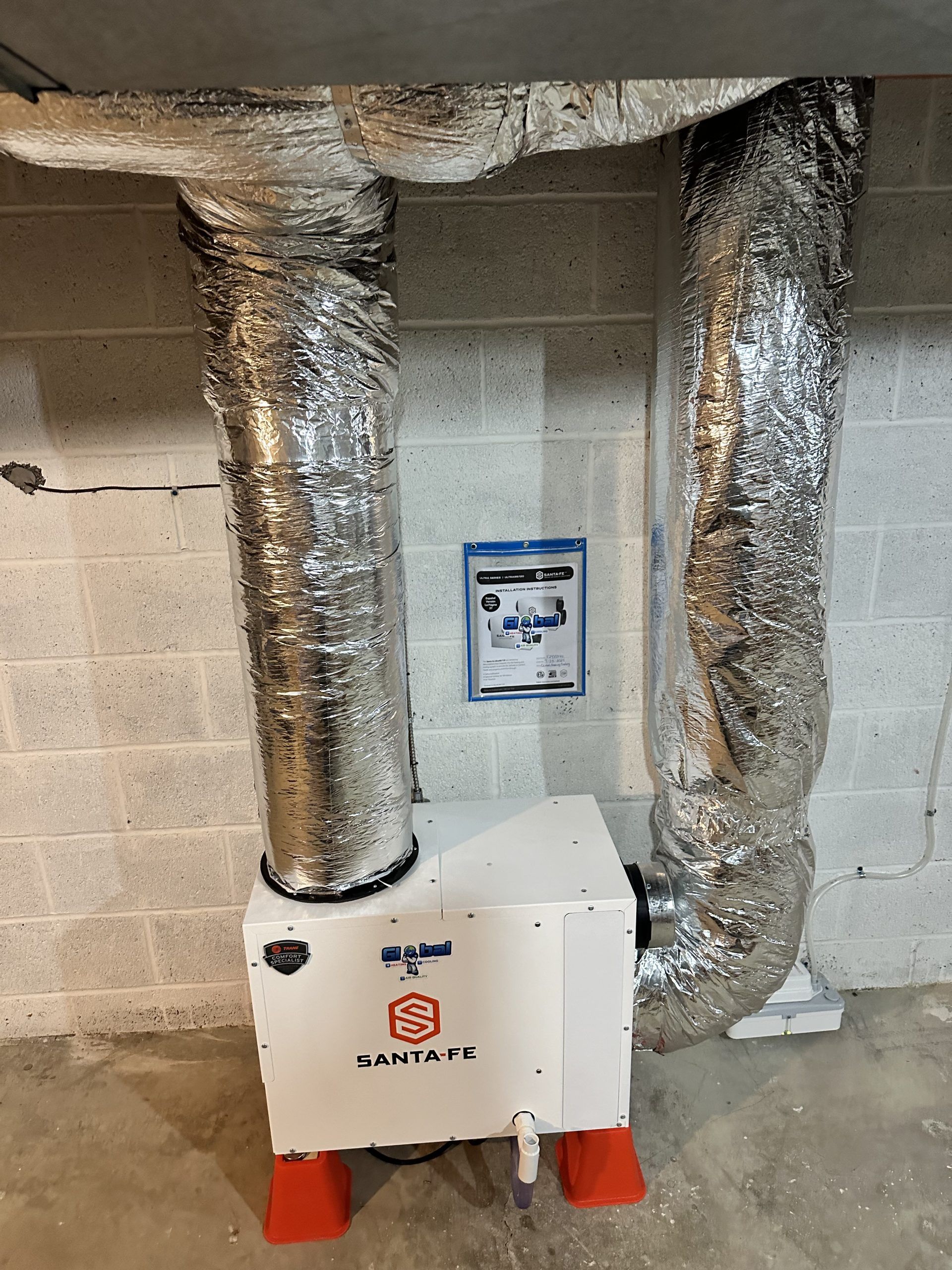 White air purifier unit with silver ducting, against a cinder block wall.
