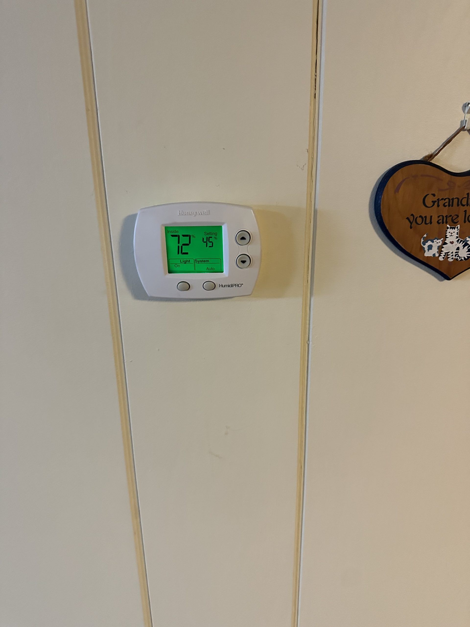 White thermostat on a beige wall, displaying the temperature in green; a wooden heart decoration is to the right.