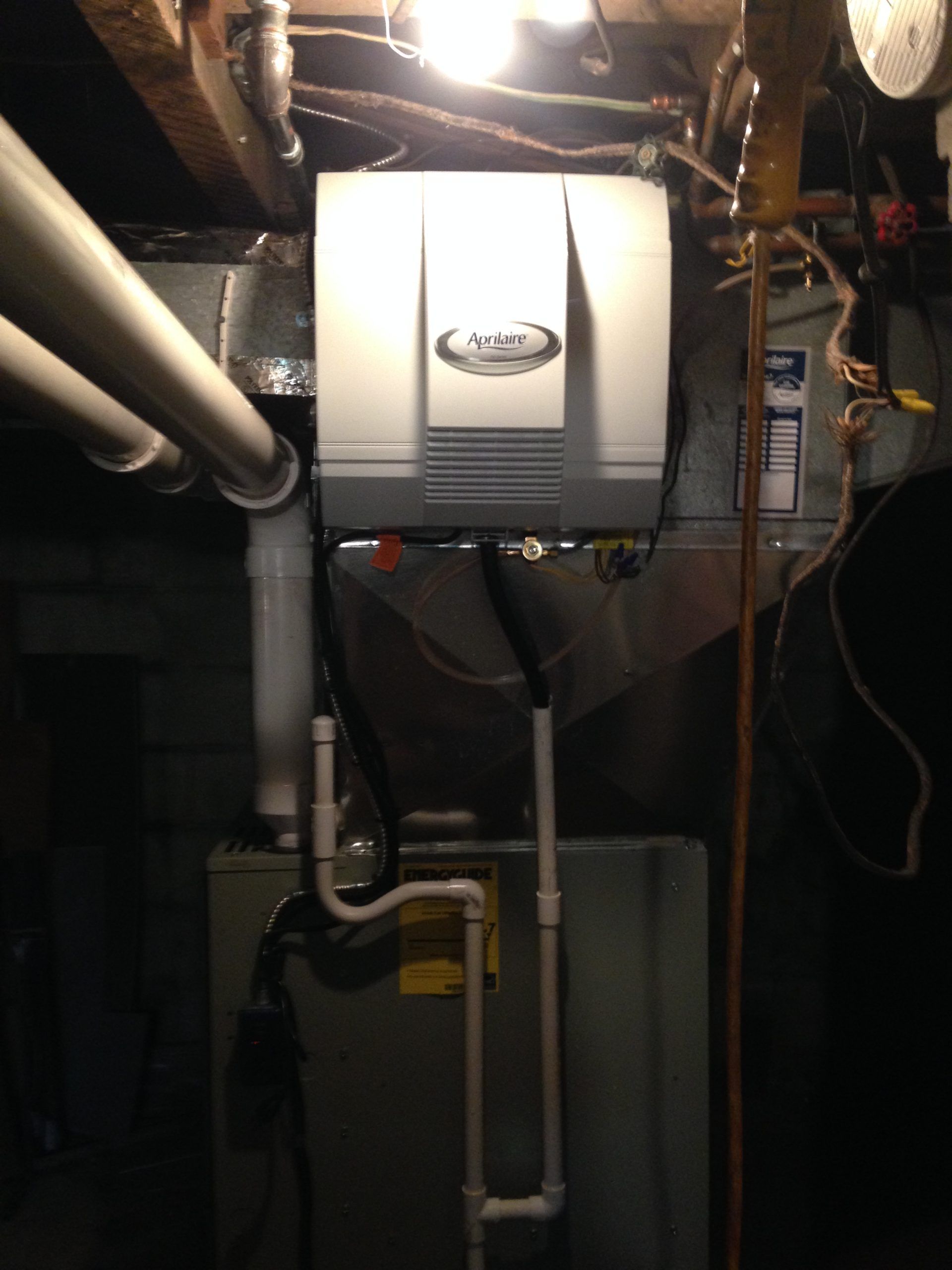 Furnace and humidifier unit in a basement setting. White humidifier atop a gray metal furnace. Pipes and wires are visible.