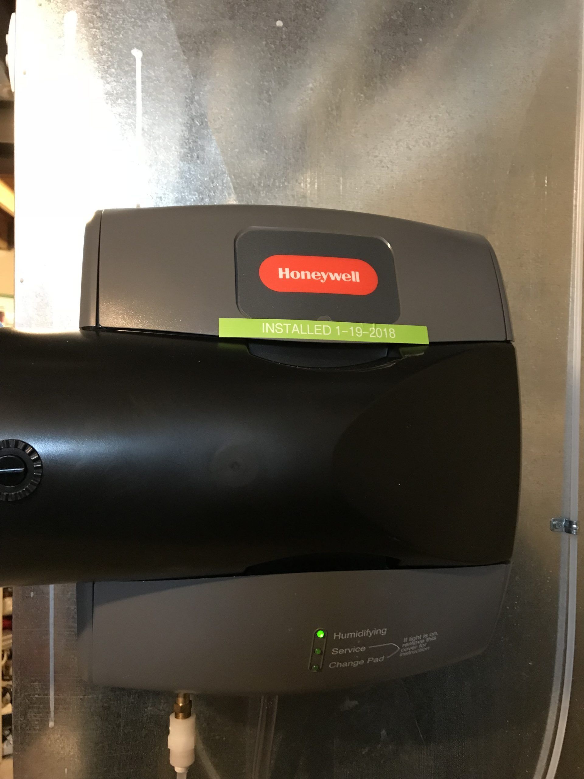 Honeywell humidifier attached to metal ductwork, dark gray and black, with a green power light lit.