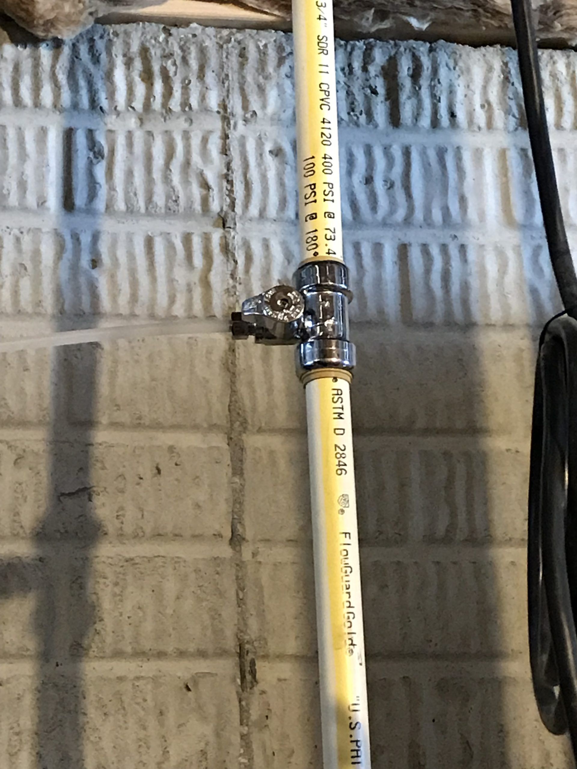 Vertical yellow PVC pipe with a chrome-colored tee fitting and valve on a white brick wall.