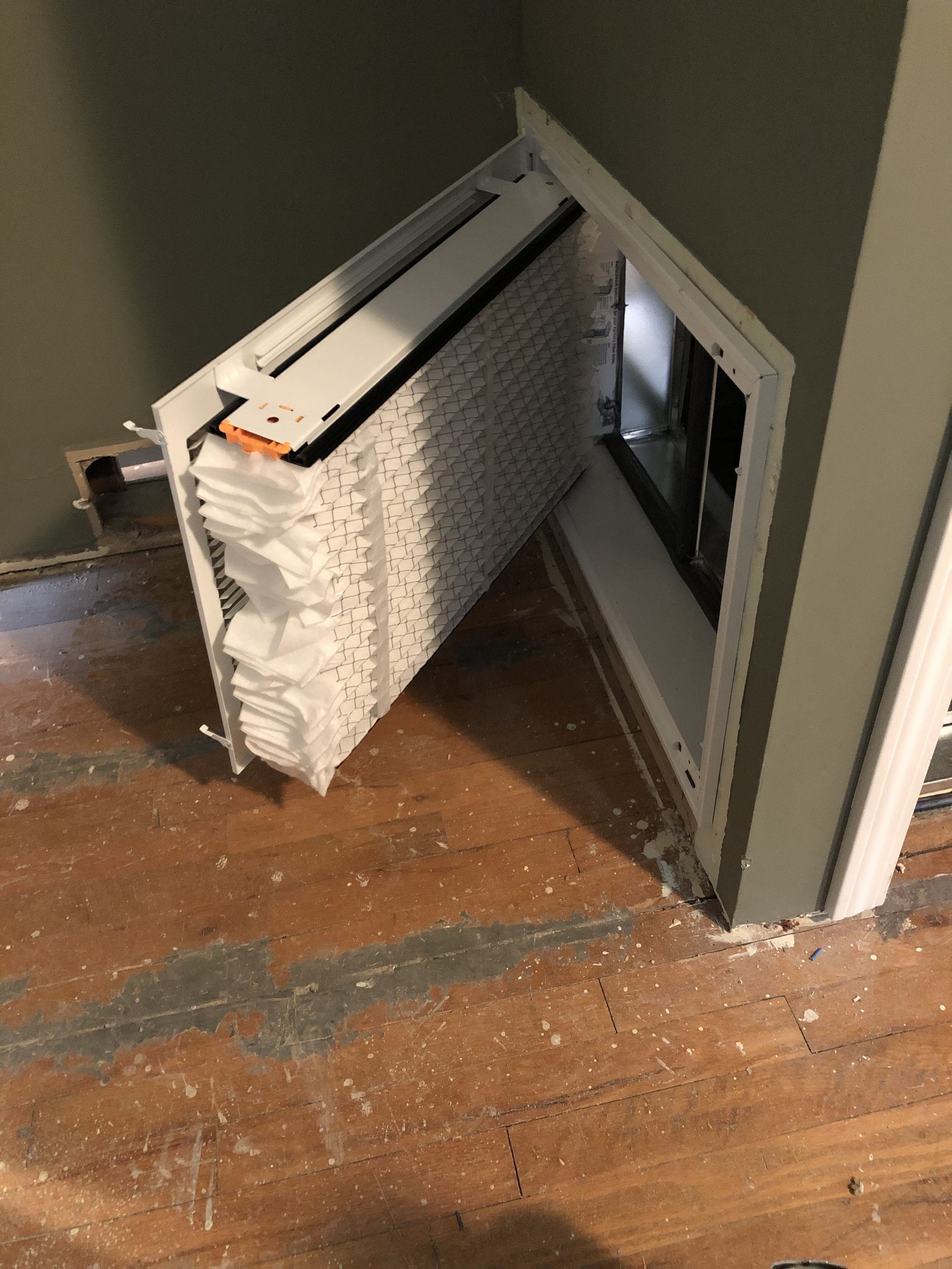 Open white vent cover revealing layered white filter, in a doorway with damaged wooden flooring.