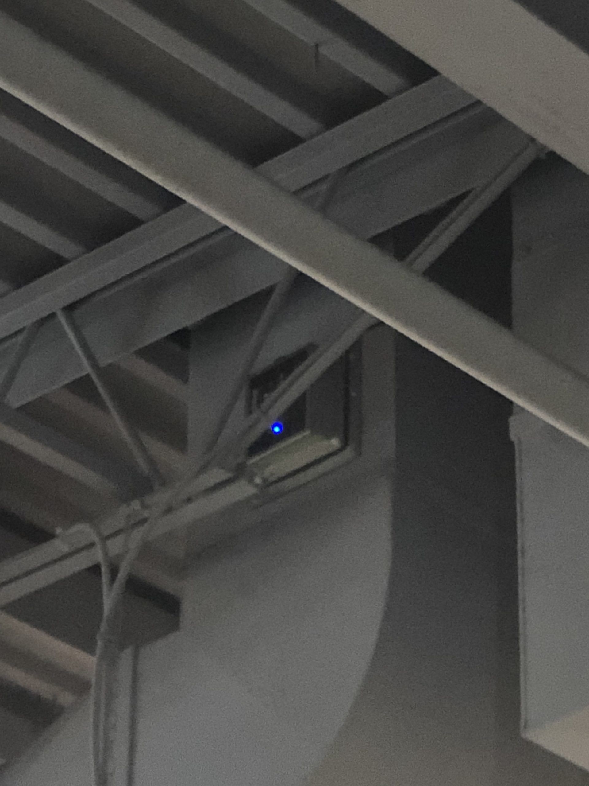 Blue light emitting from a metal box on a support beam within a gray, industrial setting.