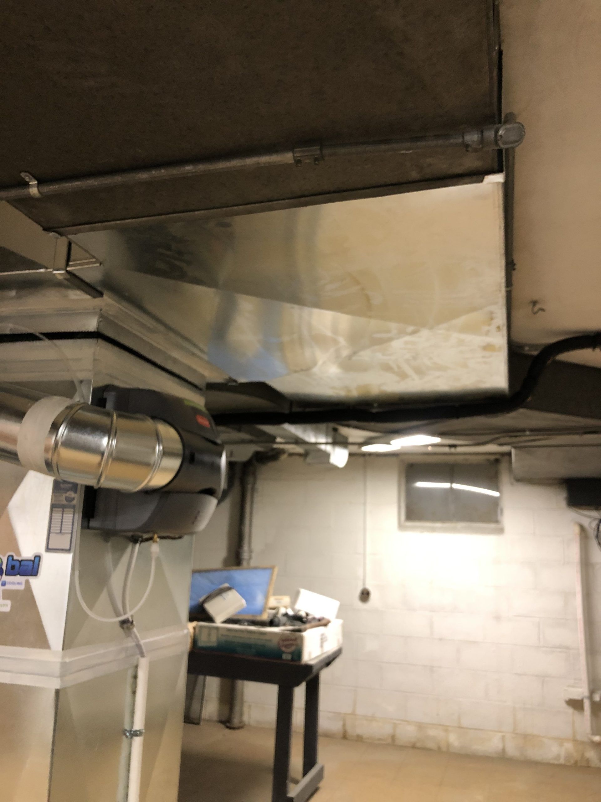 Metal ductwork installed on a basement ceiling with a small window and a white-painted brick wall.