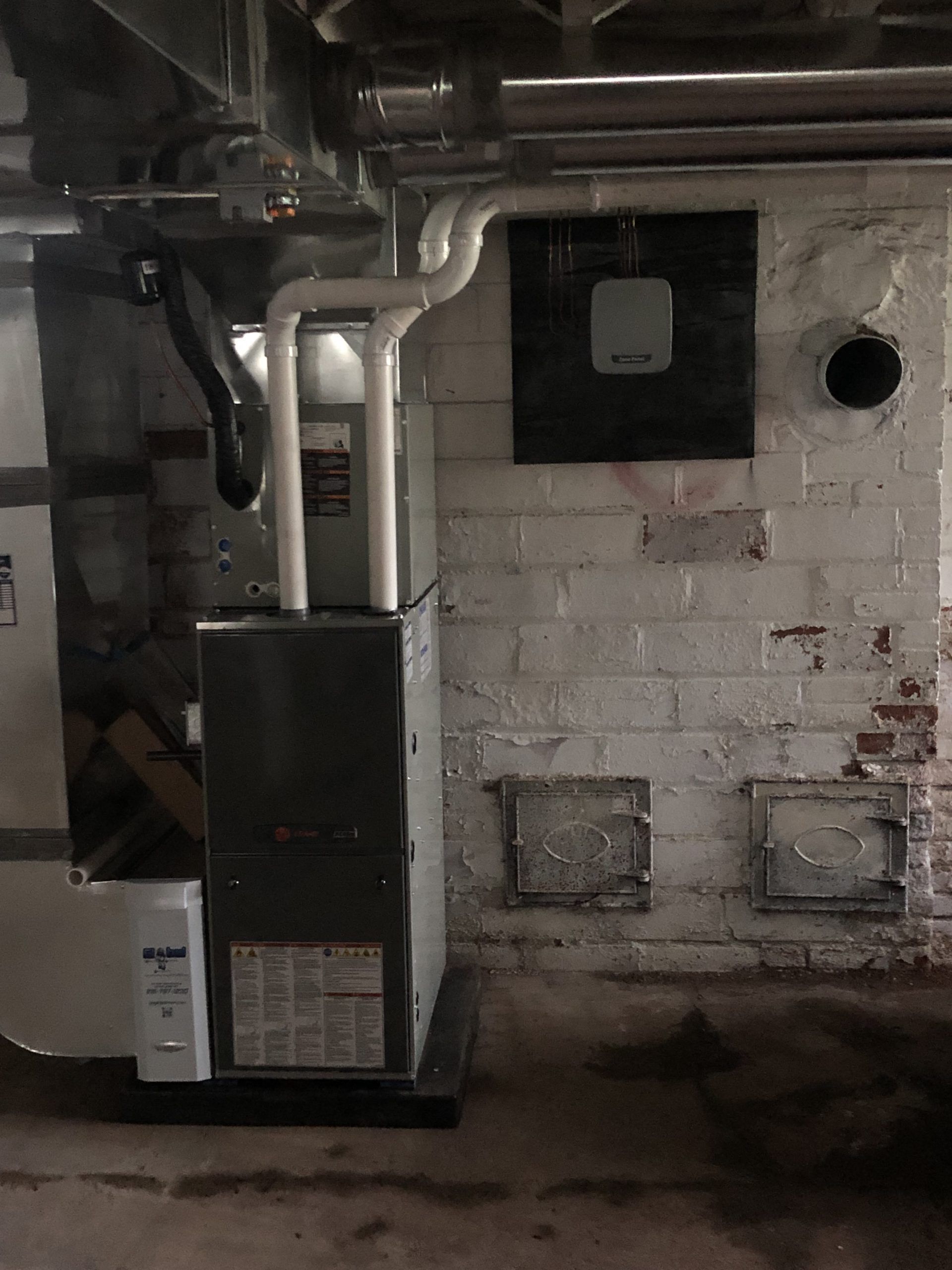 Furnace and ductwork in a basement against a brick wall.  White pipes, black and gray metal, and floor vents are visible.
