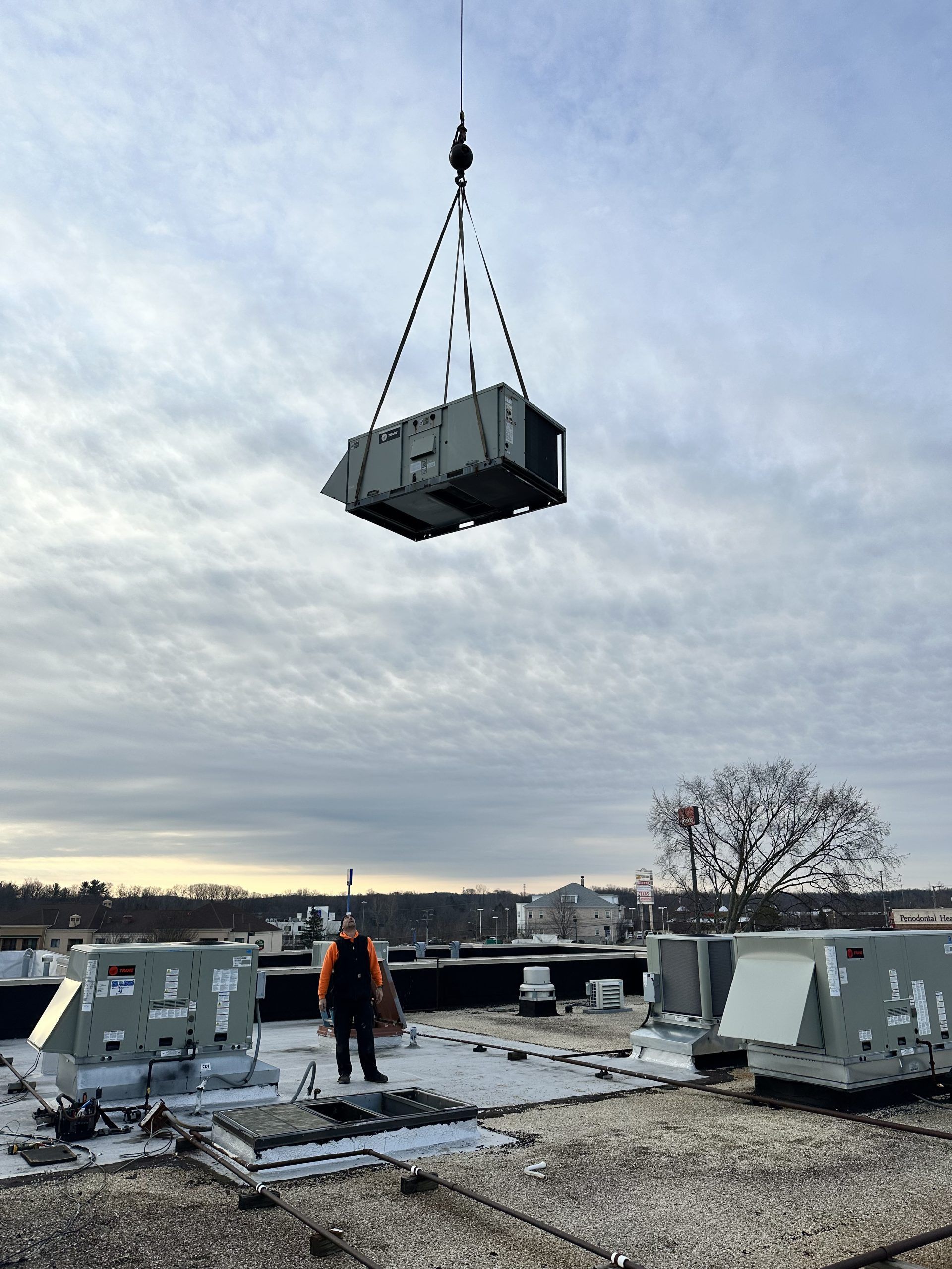 A rooftop worker watches a crane lift an HVAC unit on a cloudy day.