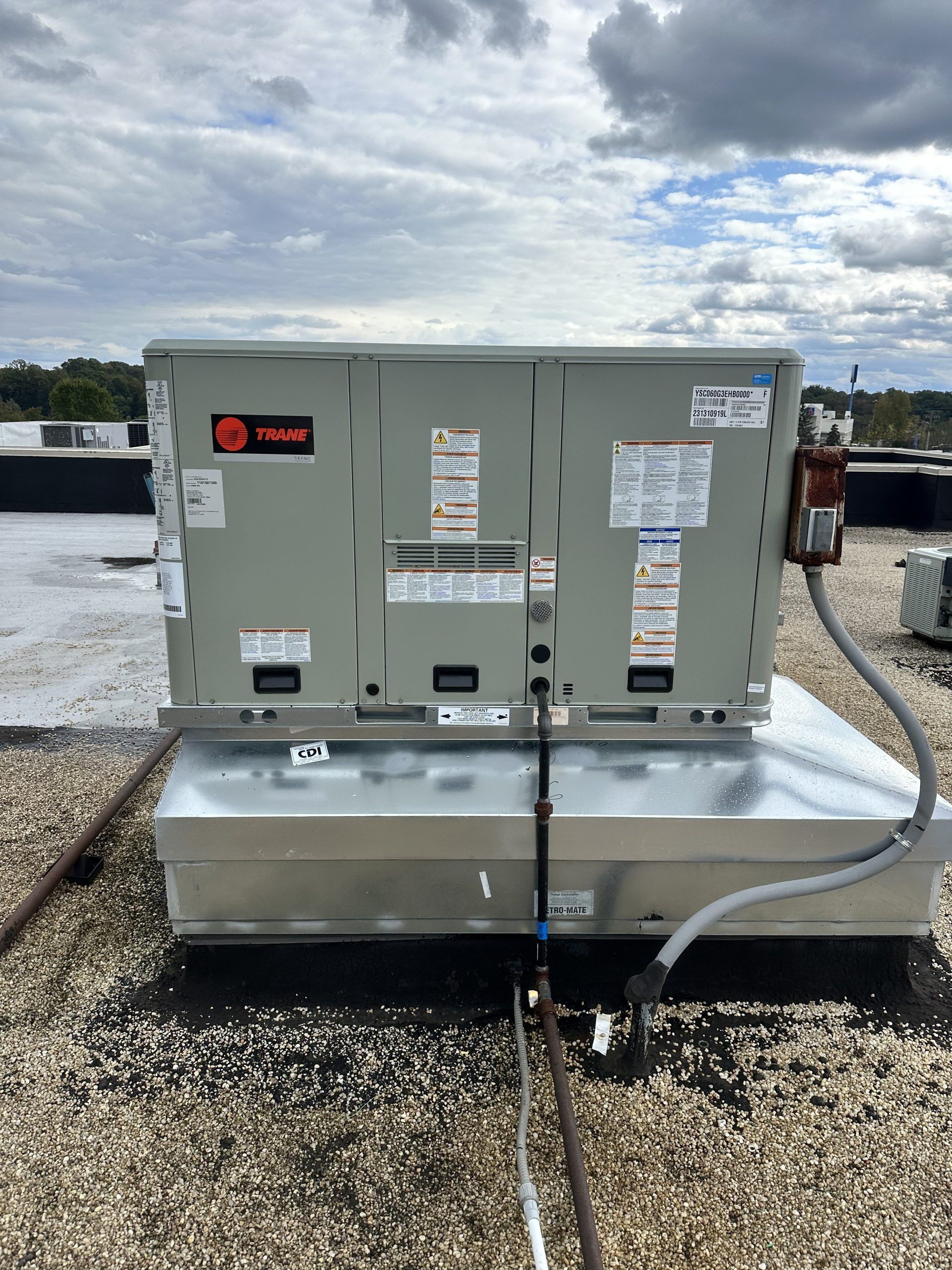 HVAC unit atop a flat roof with overcast sky. Grey unit, silver base, black roof, and conduit.