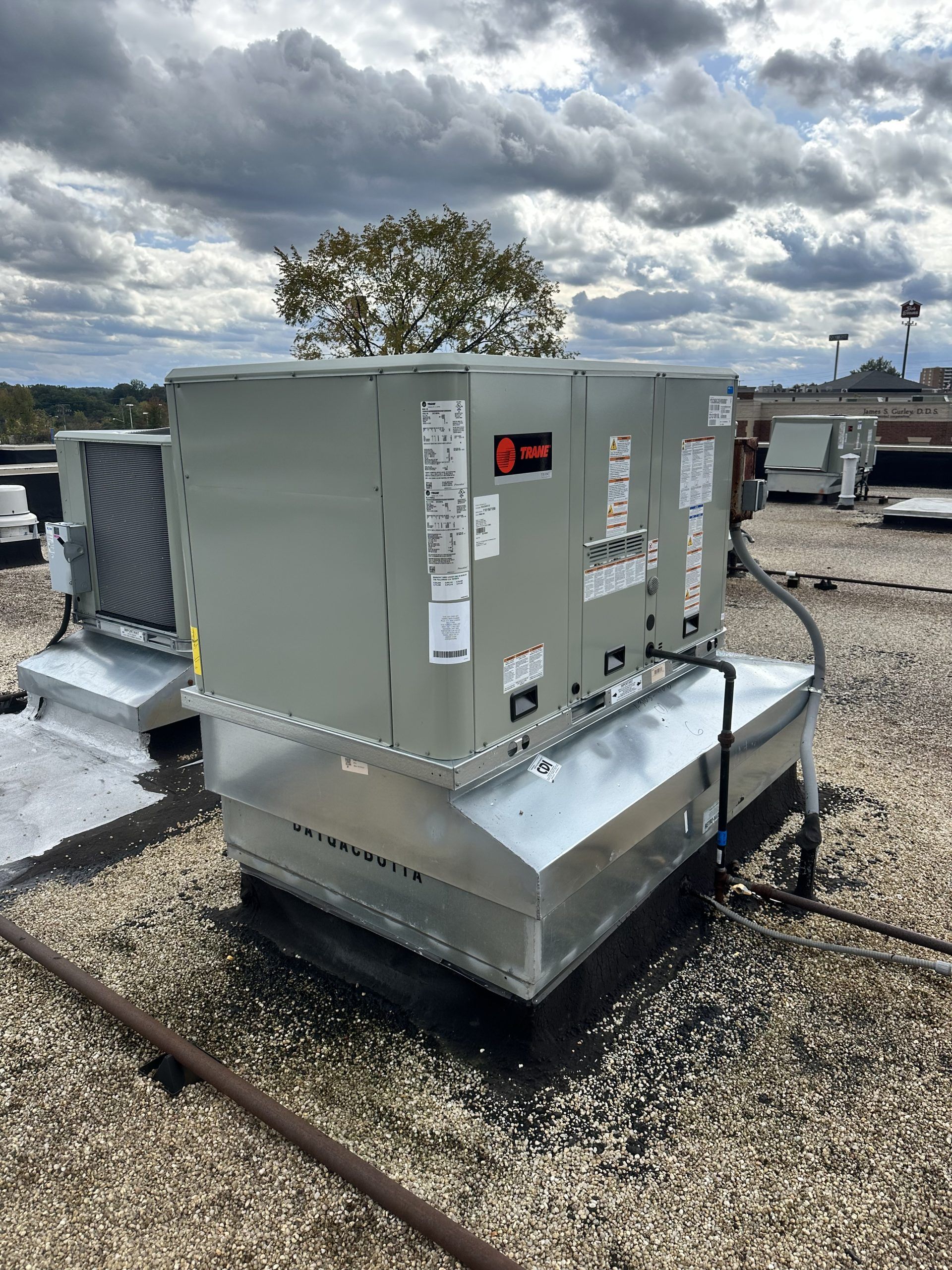 HVAC unit on a rooftop, with a cloudy sky in the background. Other units and a tree are also visible.