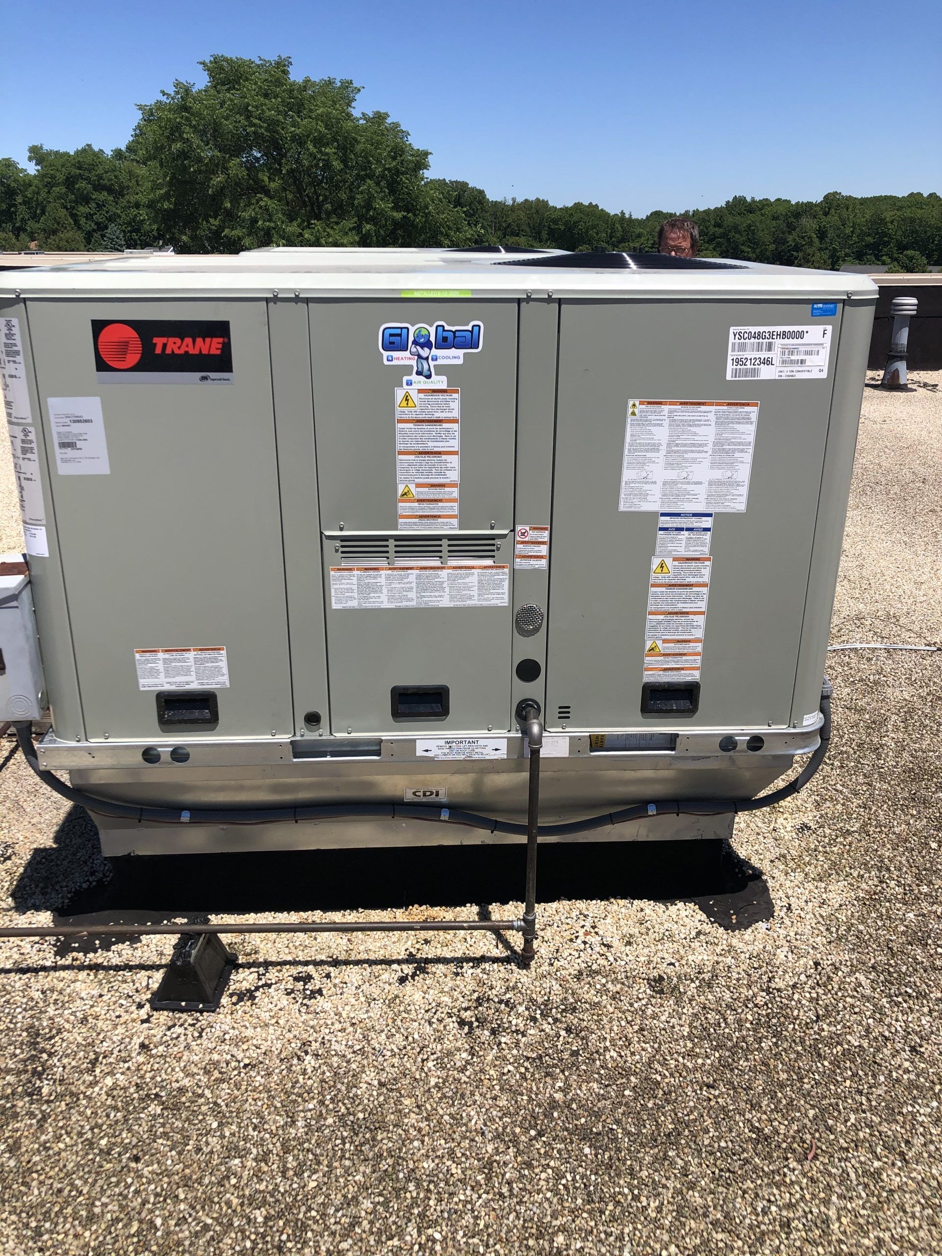 Gray rooftop HVAC unit with safety labels, Trane logo, on a flat roof under a blue sky.