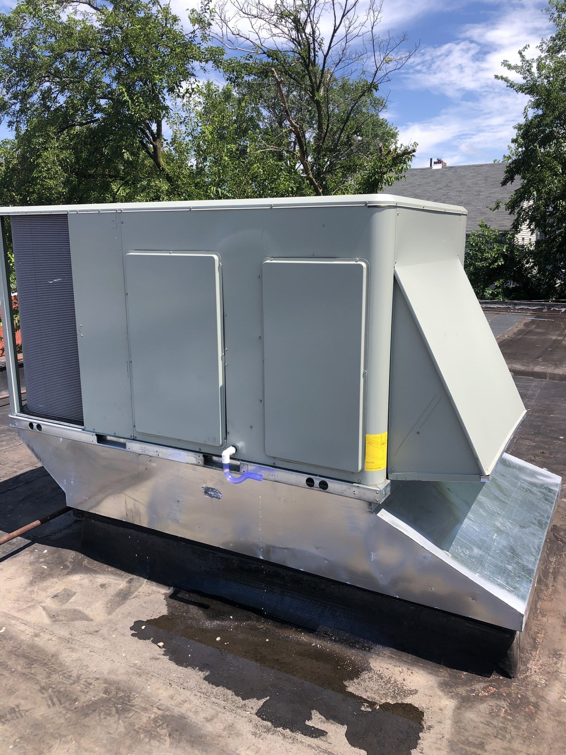Rooftop HVAC unit, gray metal, on a flat roof with trees in the background.