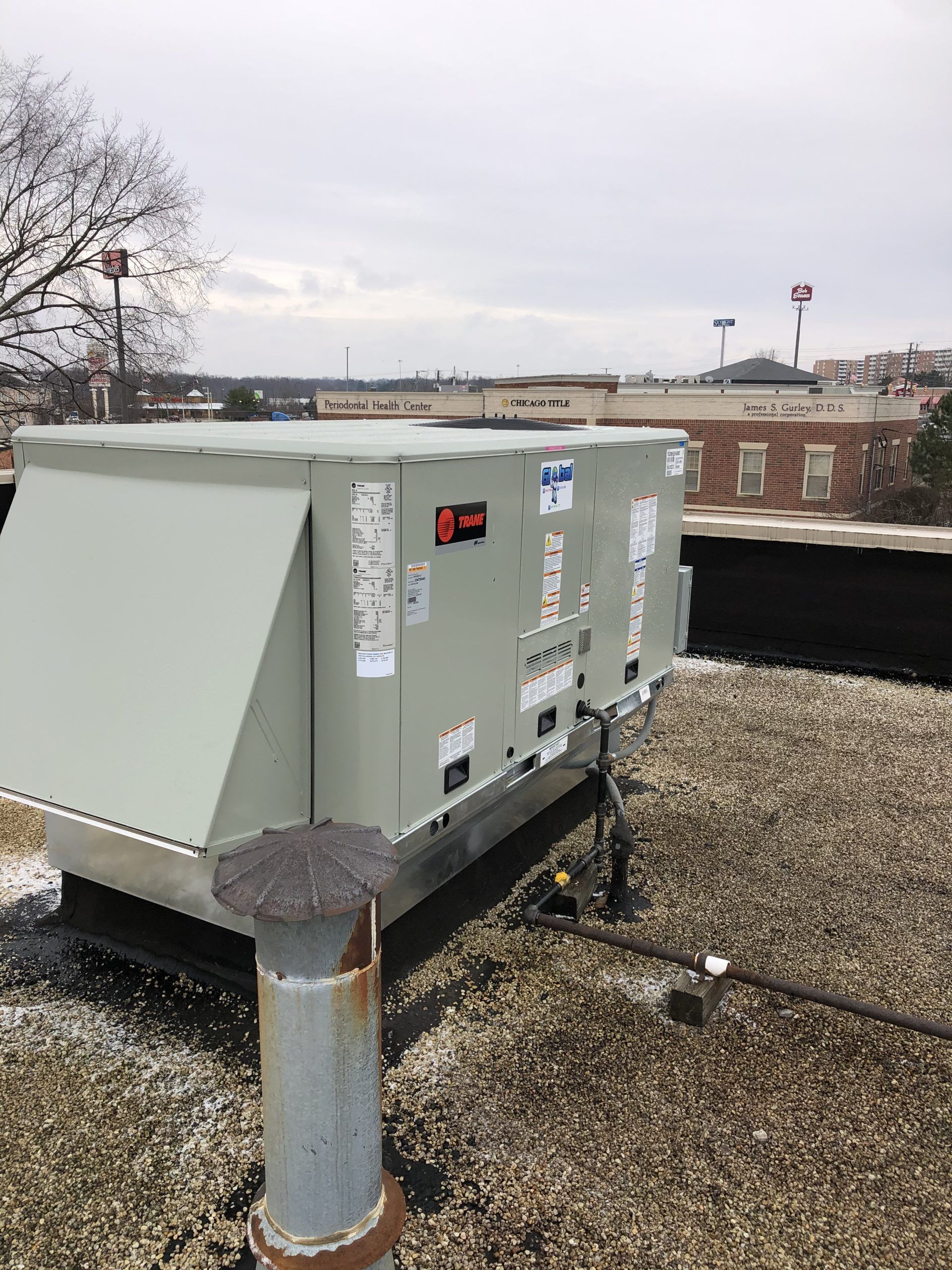Rooftop HVAC unit on a flat roof. Gray metal box with a vent, overcast sky, buildings in the background.