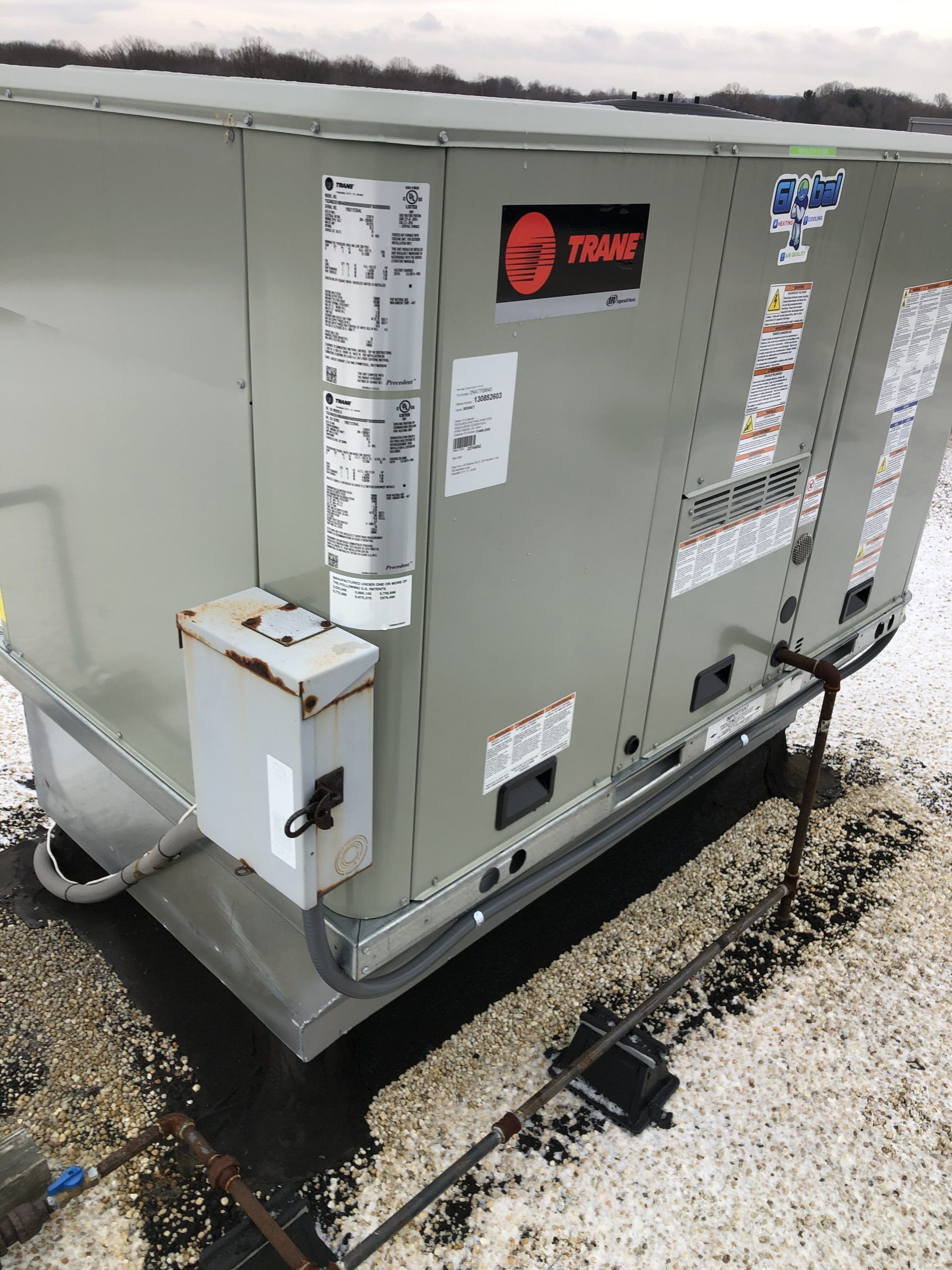 HVAC unit on a rooftop with Trane logo and electrical box. Gray metal exterior with labels and pipes.