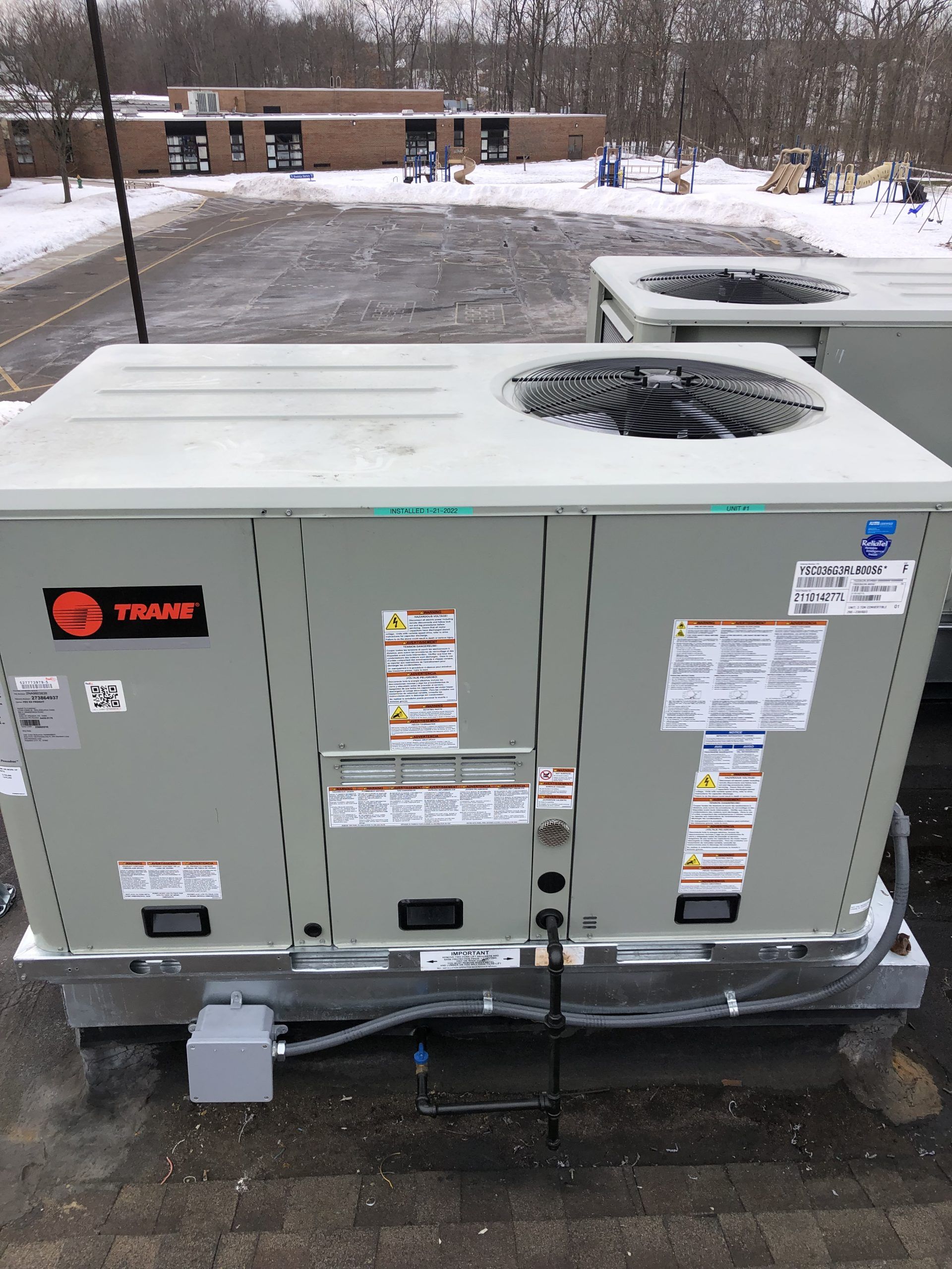 A rooftop HVAC unit covered in snow on a cloudy day. Other units and a building are in the background.