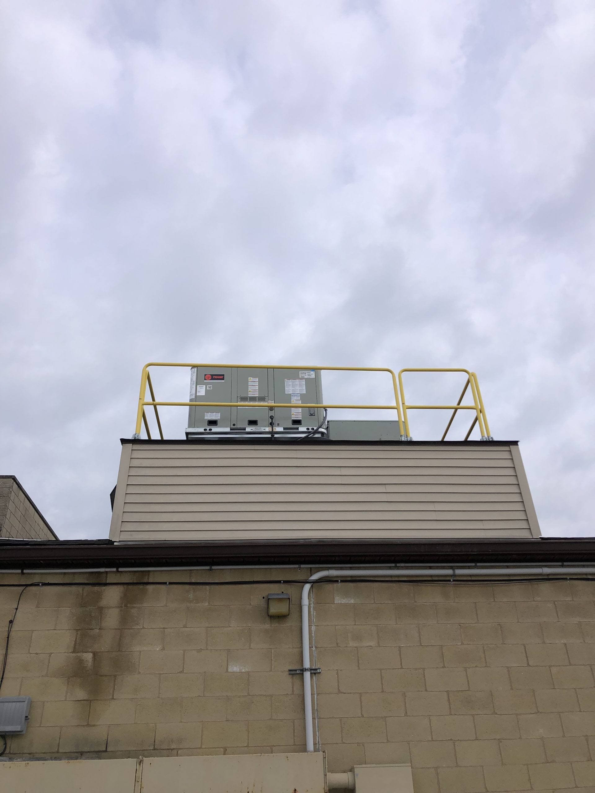 HVAC unit on a roof with yellow safety railing. Overcast sky. Beige and tan brick building exterior.