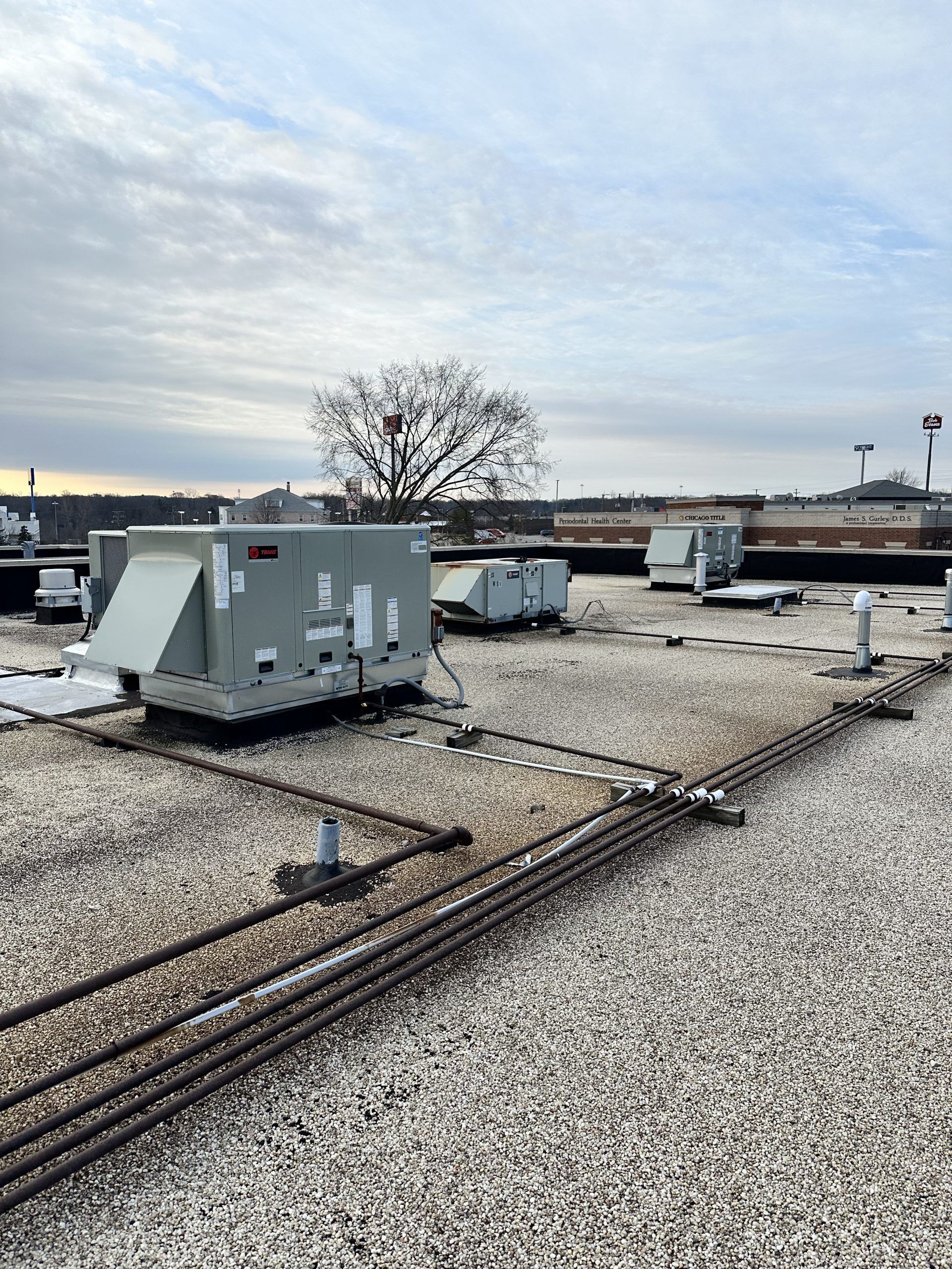 Rooftop with HVAC units and pipes against a cloudy sky, gravel surface. A tree in the distance.