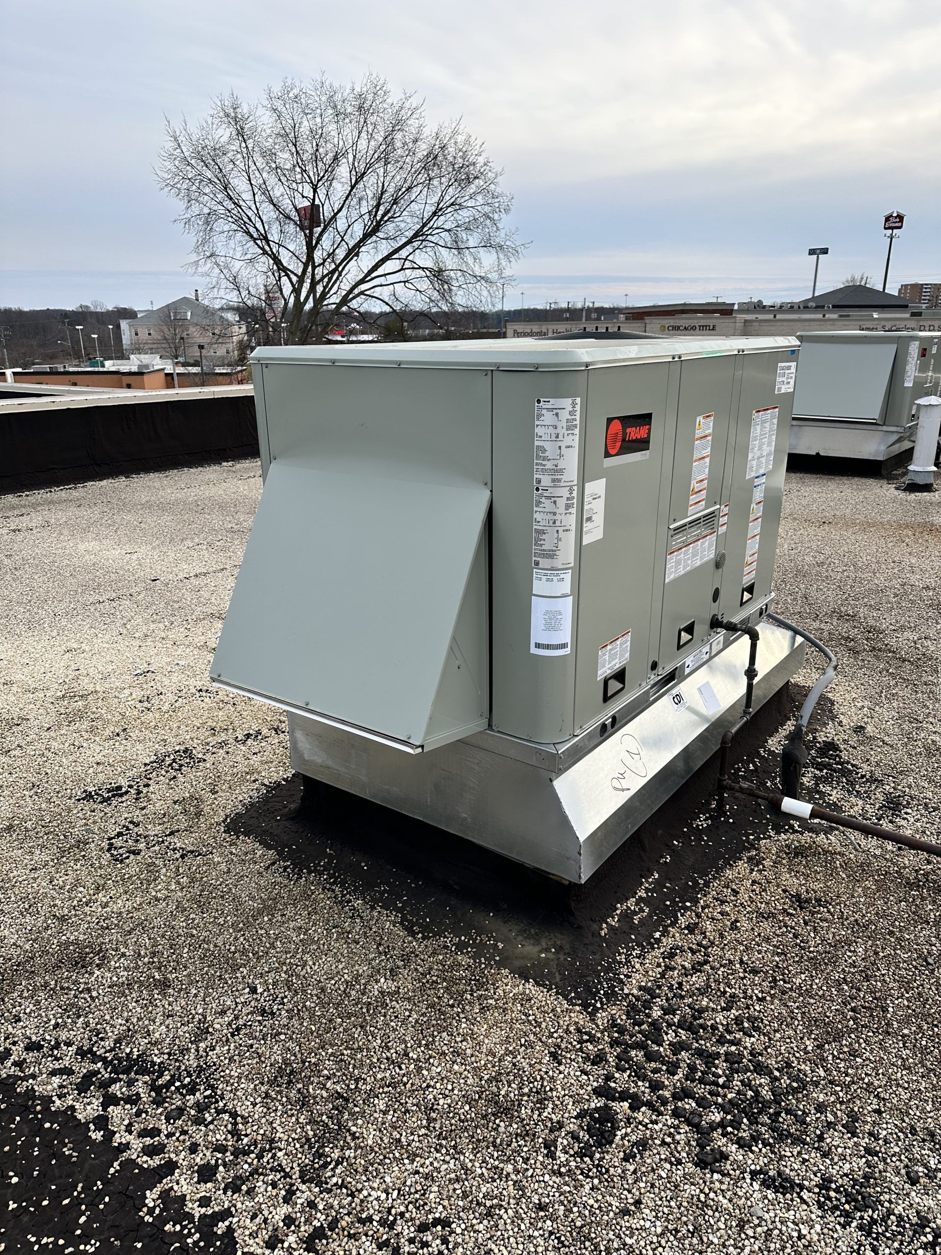 Rooftop HVAC unit on a flat roof with a tree and other units in the background. Gray and silver colors.
