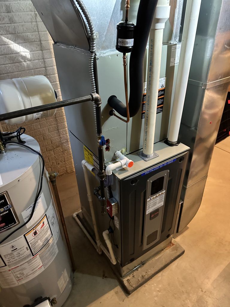 Furnace and water heater in a basement. The furnace is gray and rectangular, with pipes and a black filter. 