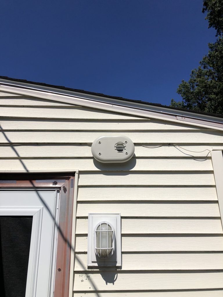 A light tan alarm system and a white exterior light on the side of a house, against a blue sky.