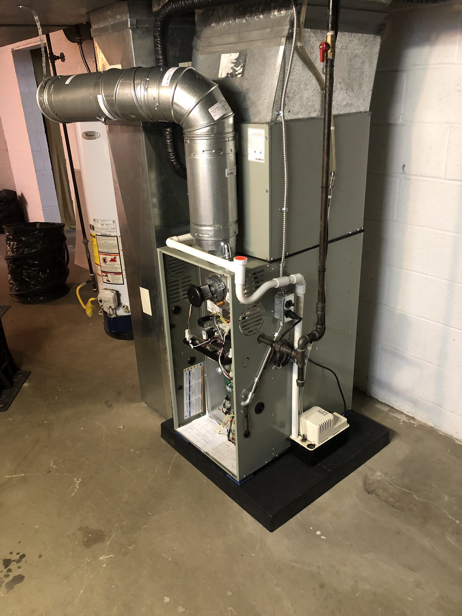 Furnace unit in a basement, with silver ductwork, pipes, and electrical components, against a white wall.