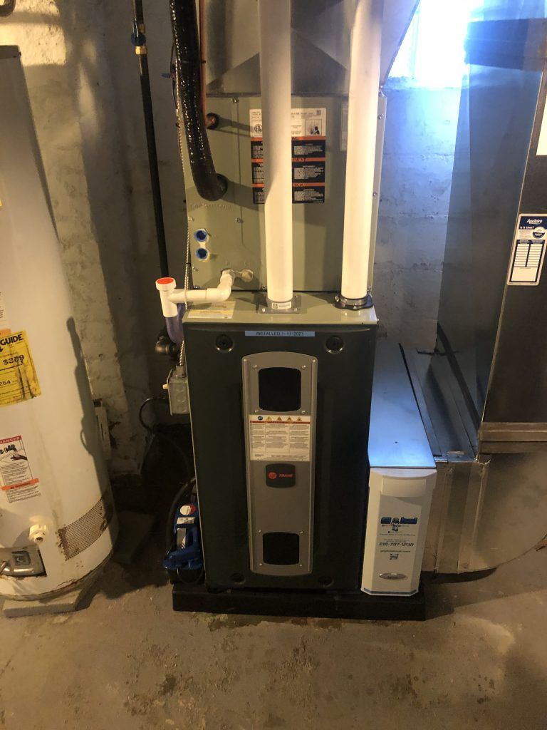 Furnace in a basement; gray metal unit with white pipes and air filter. Water heater to the left.