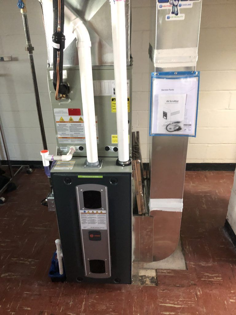 A furnace with two white PVC pipes and a metal vent pipe. A document in a plastic sleeve is on the wall.